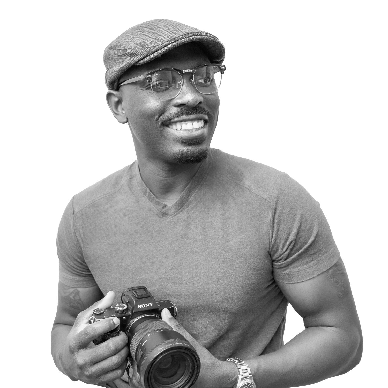 Professional photographer wearing glasses and a newsboy cap holds a DSLR camera while smiling in black and white portrait.
