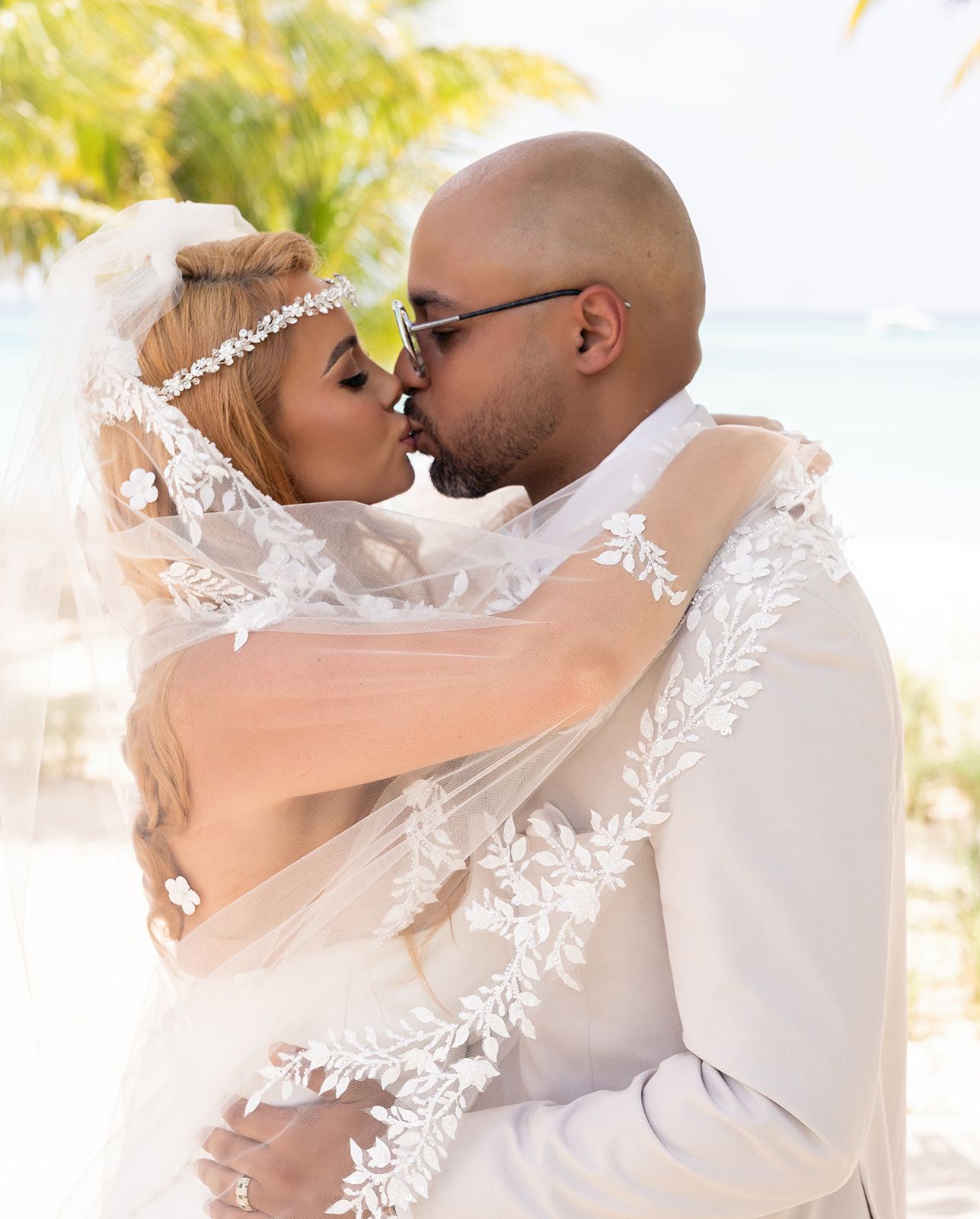 A loving couple shares a romantic kiss at their beach wedding, with delicate lace veil details and tropical palm trees in background.