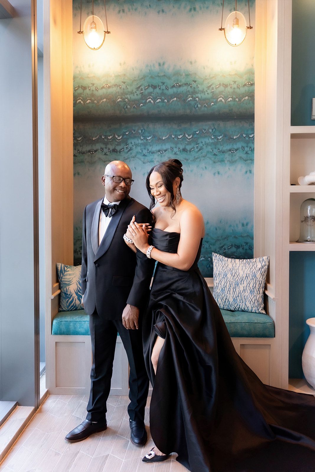 A couple in elegant black formal wear share an intimate moment in a room with turquoise wall art.