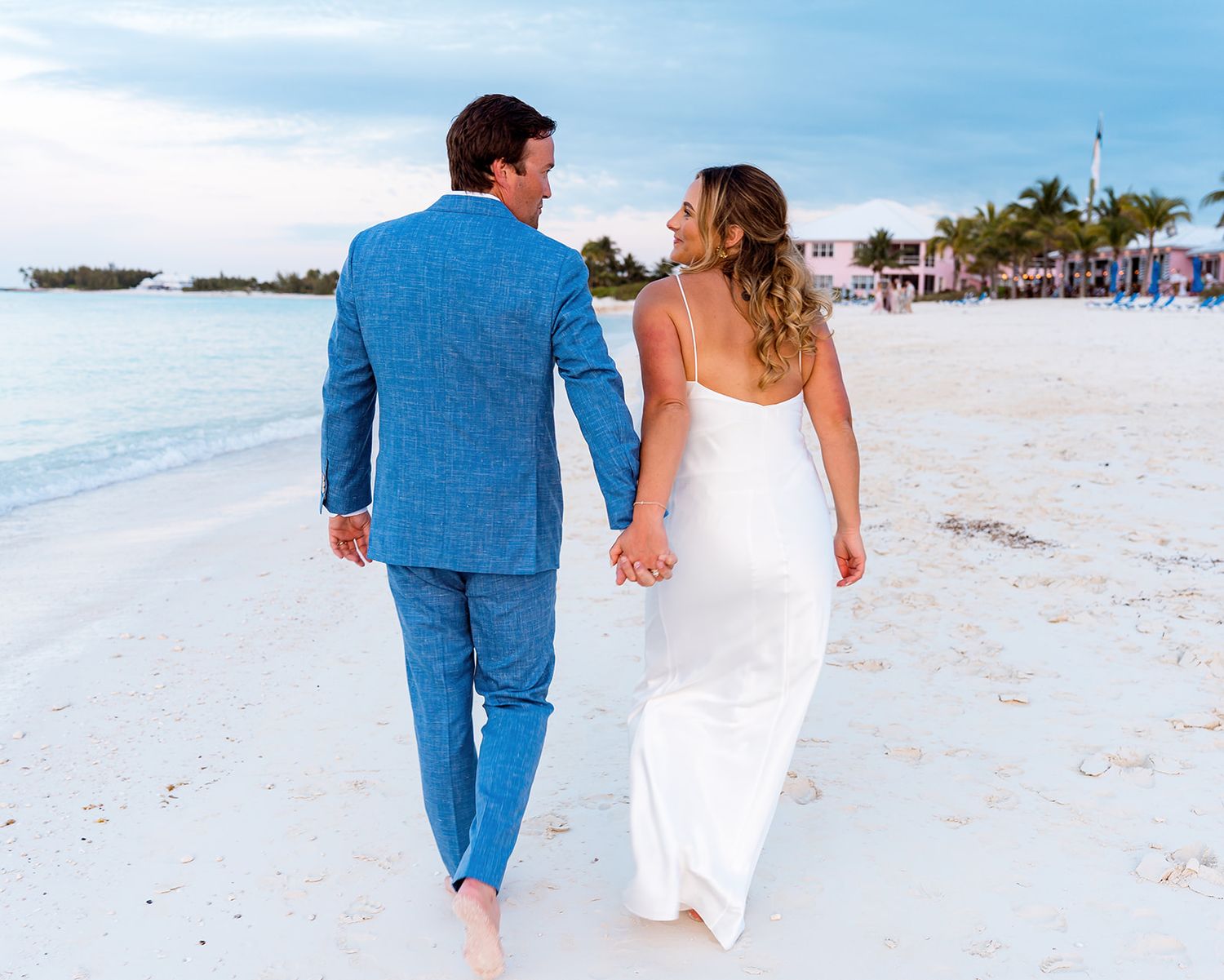 Couple strolling barefoot on tropical beach with bright blue suit and white wedding dress.