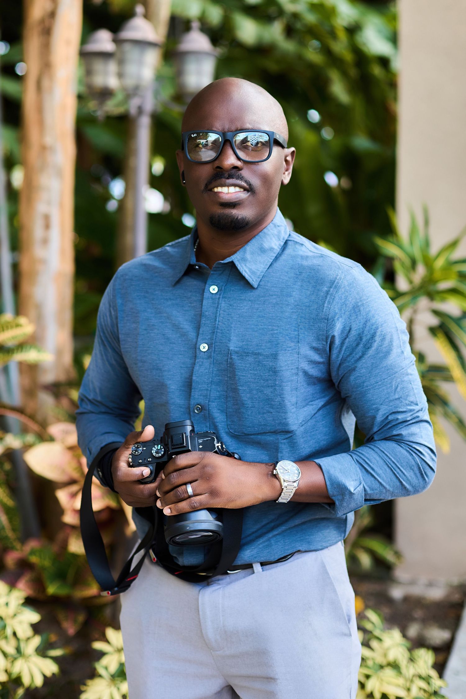 Professional photographer wearing glasses and a blue button-down shirt holds a DSLR camera in an outdoor garden setting.