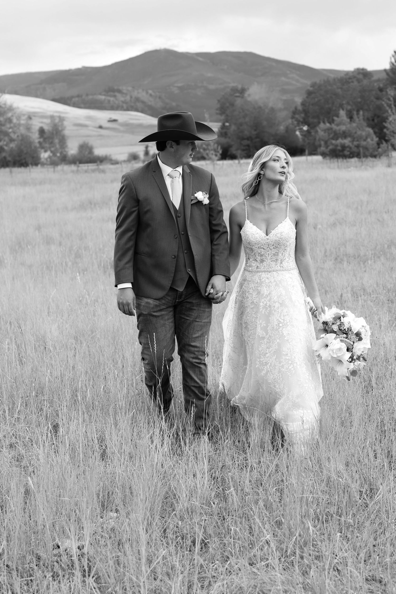 A bride in a lace gown and groom in western attire walk hand in hand through a sunny mountain meadow at their Montana summer wedding.