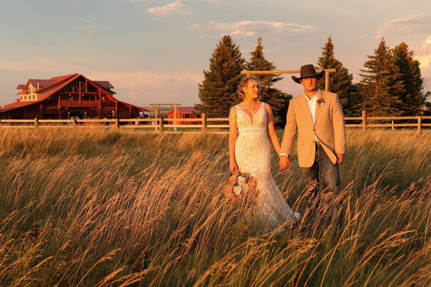 A rustic barn wedding scene at sunset with a couple walking through golden wheat fields and tall evergreen trees.