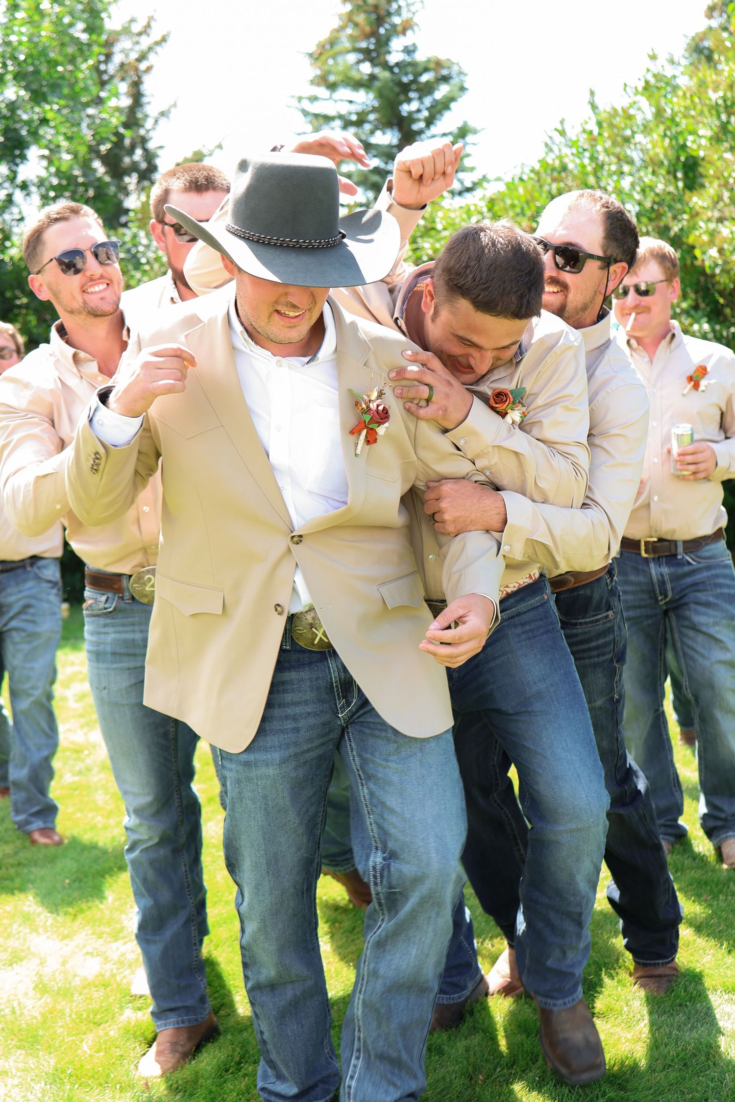 A group of groomsmen in tan vests and jeans rough up the groom on green grass at a Montana wedding.