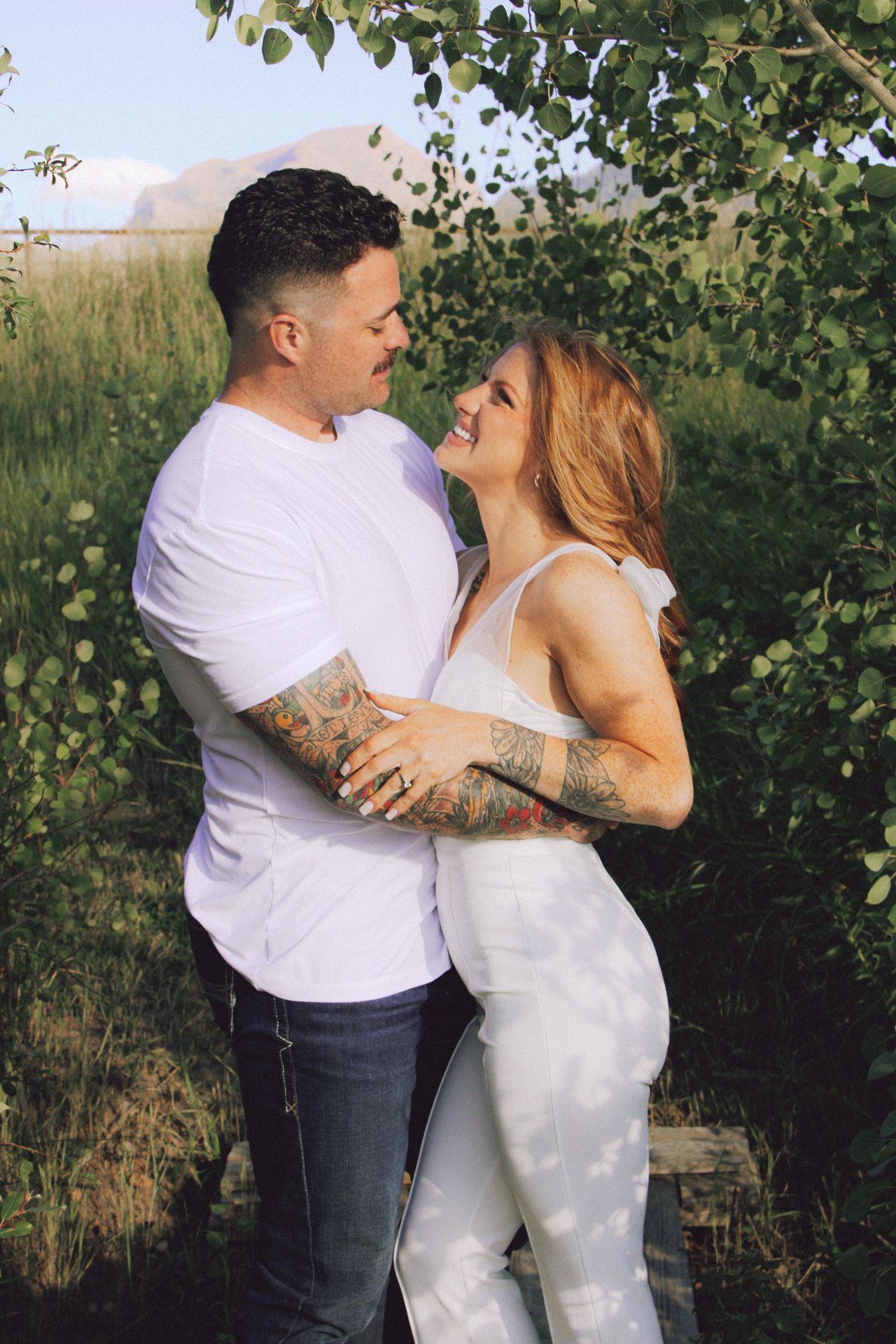 A romantic couple embraces outdoors in summer, wearing white clothing against a natural backdrop of trees, grass and Montana's mountains.