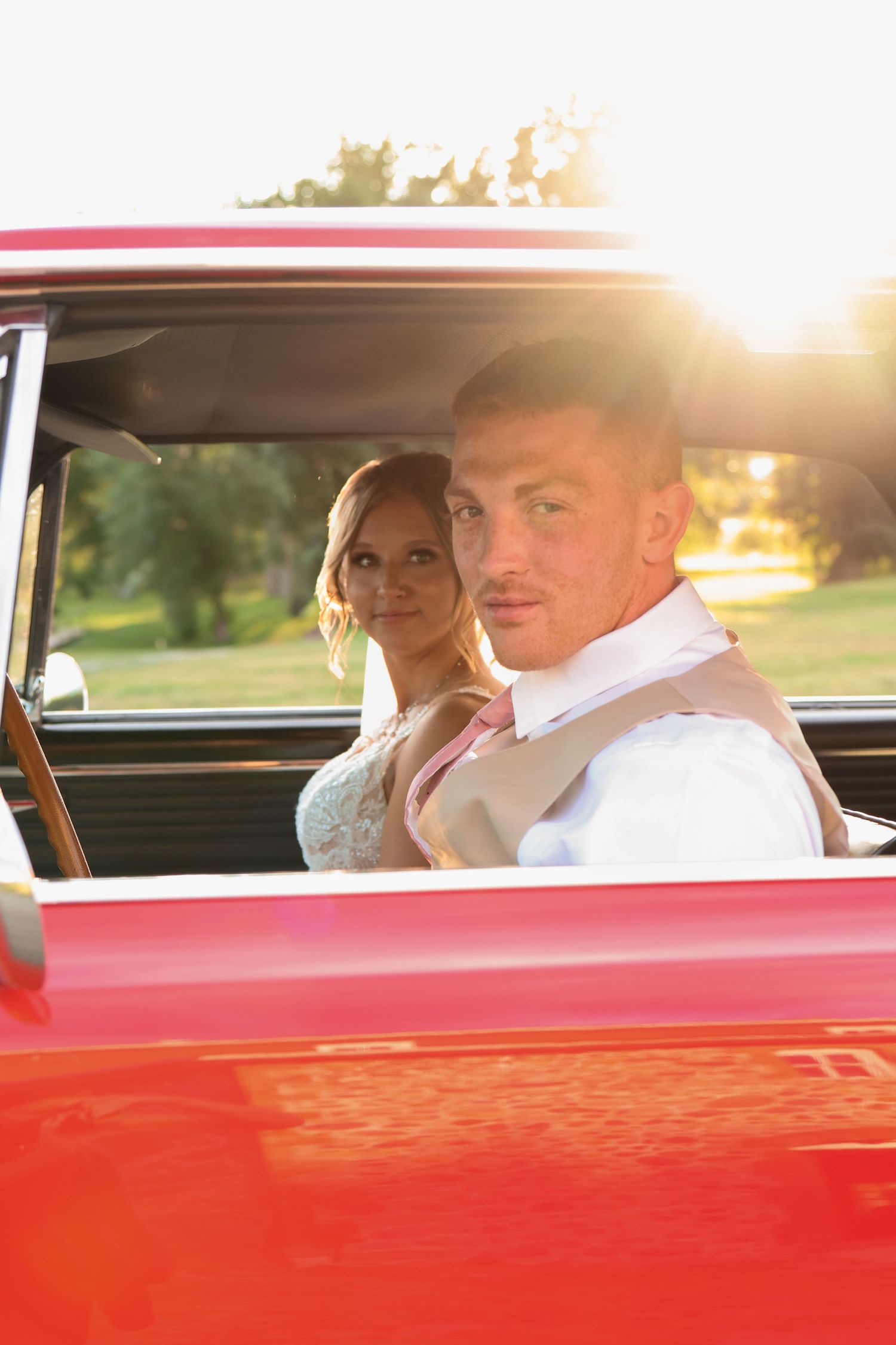 A bride and groom sits in a bright red classic car during golden hour sunset with sunlight streaming through the window.