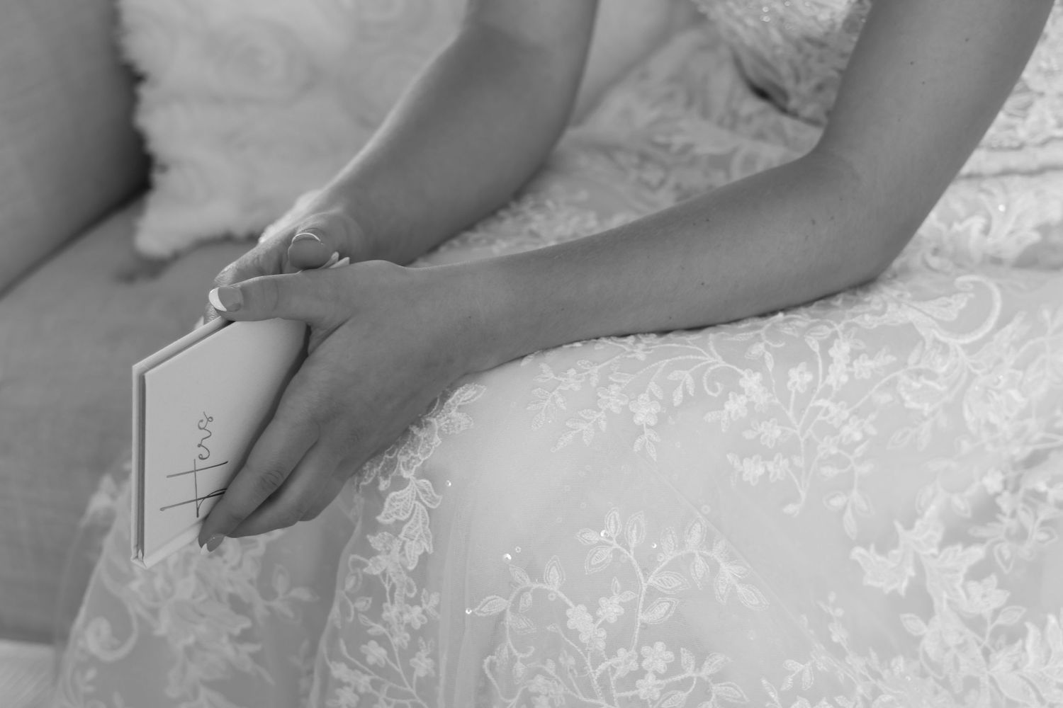 A black and white image showing hands gently holding a vow book while wearing a lace wedding dress.