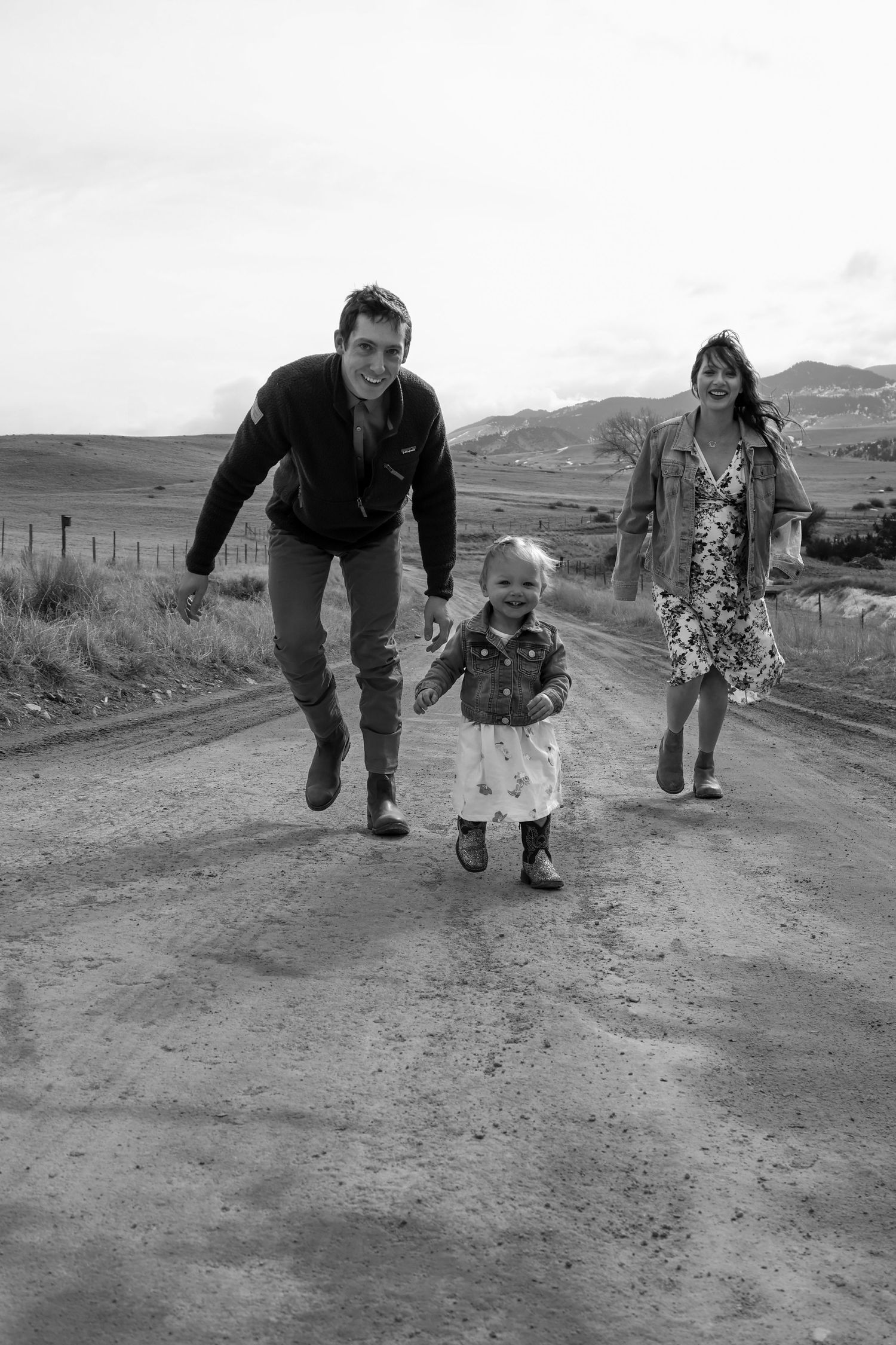 A family walks together on a rural dirt road with rolling hills in the background, captured in black and white.