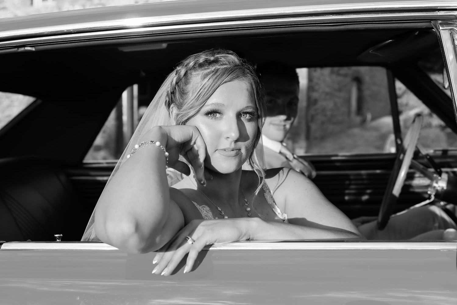 A smiling woman in a bridal veil leans out of a vintage car window in a classic black and white photograph.