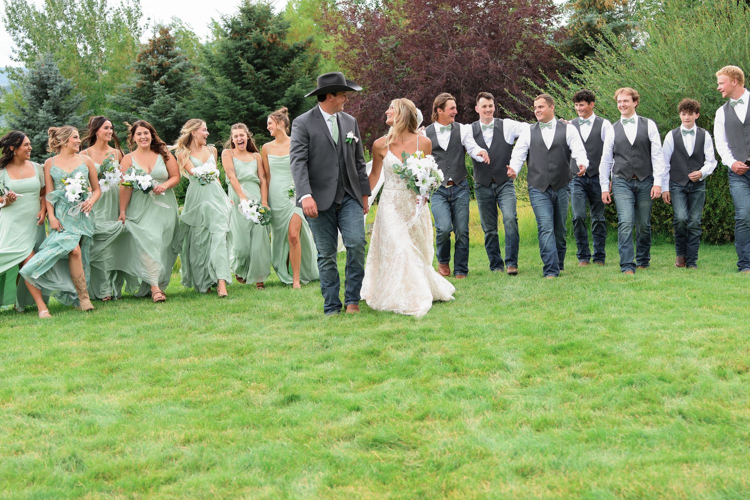 Wedding party in sage green dresses and gray vests walks together on a grassy lawn during outdoor Montana wedding in the summer.