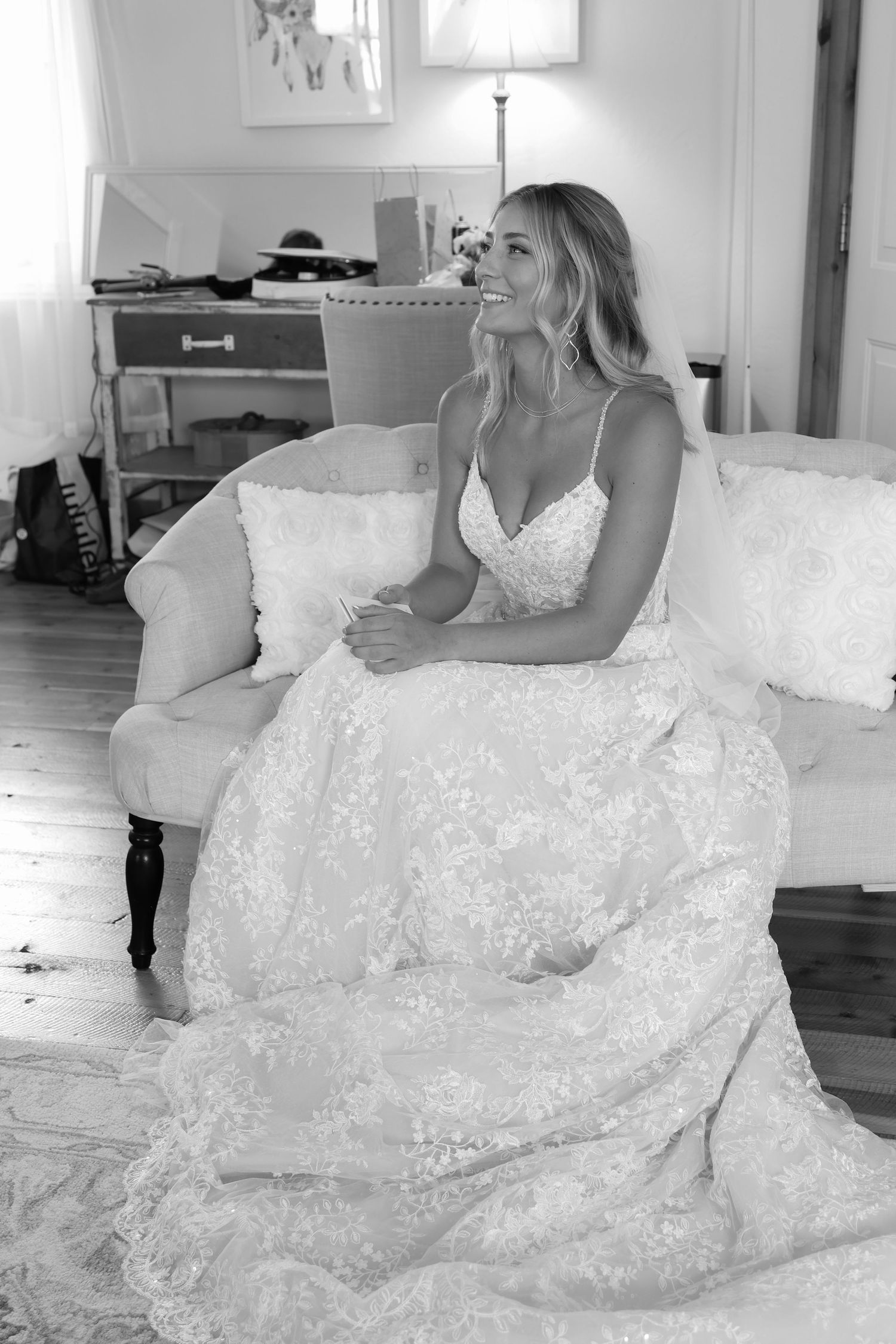 Black and white photo showing woman in elegant lace wedding gown sitting on white couch in sunlit room.