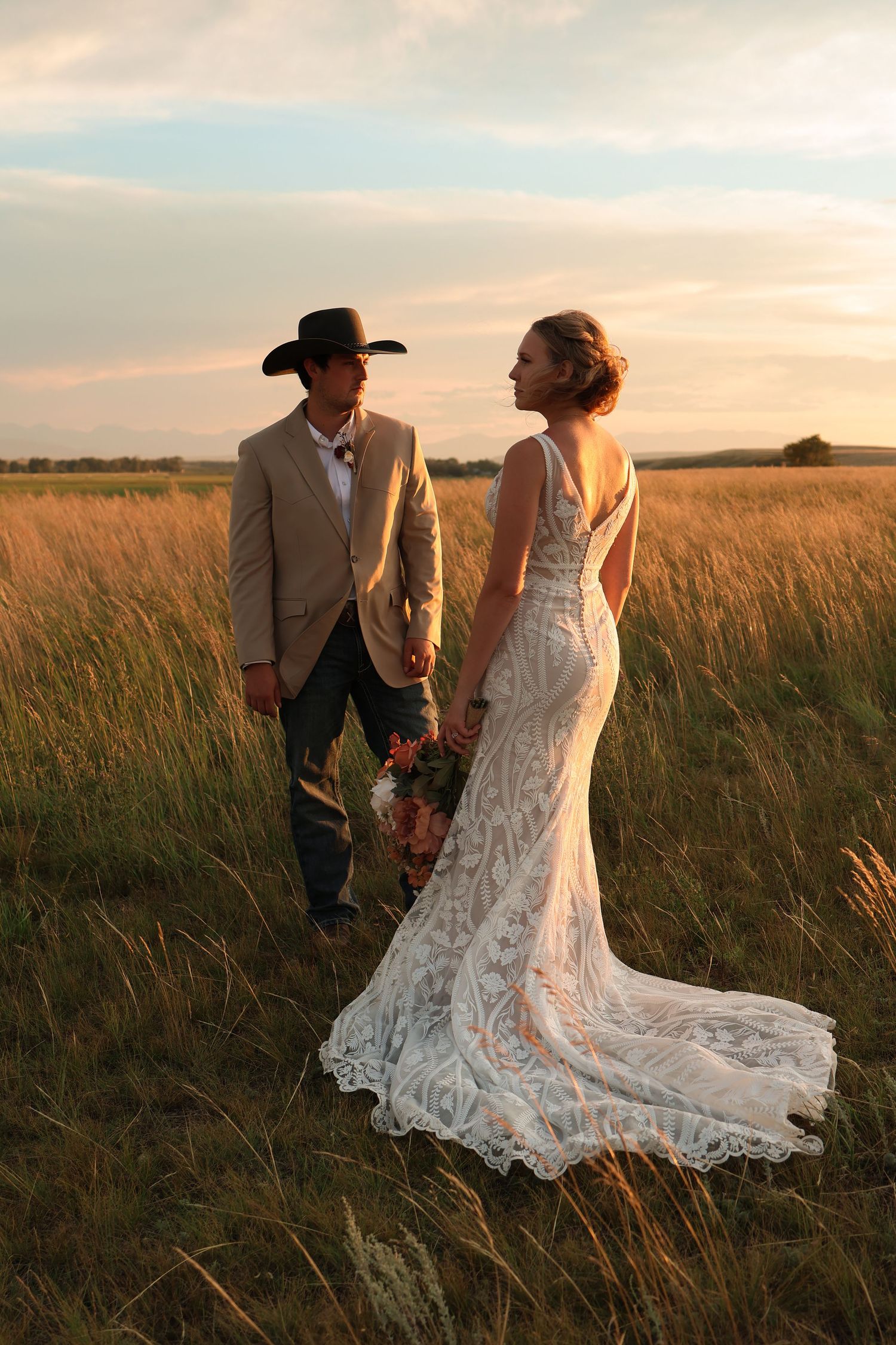 A couple in wedding attire walk through a golden wheat field at sunset during their rustic outdoor wedding photoshoot.