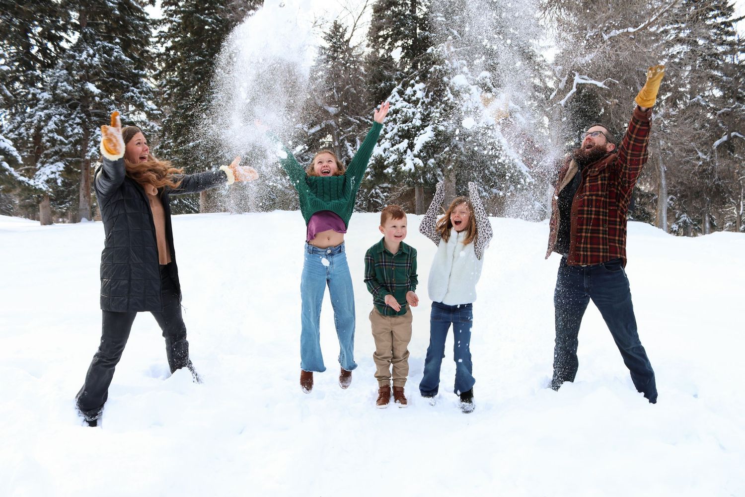 People playing and jumping in the snow during winter with snow-covered evergreen trees in the background.