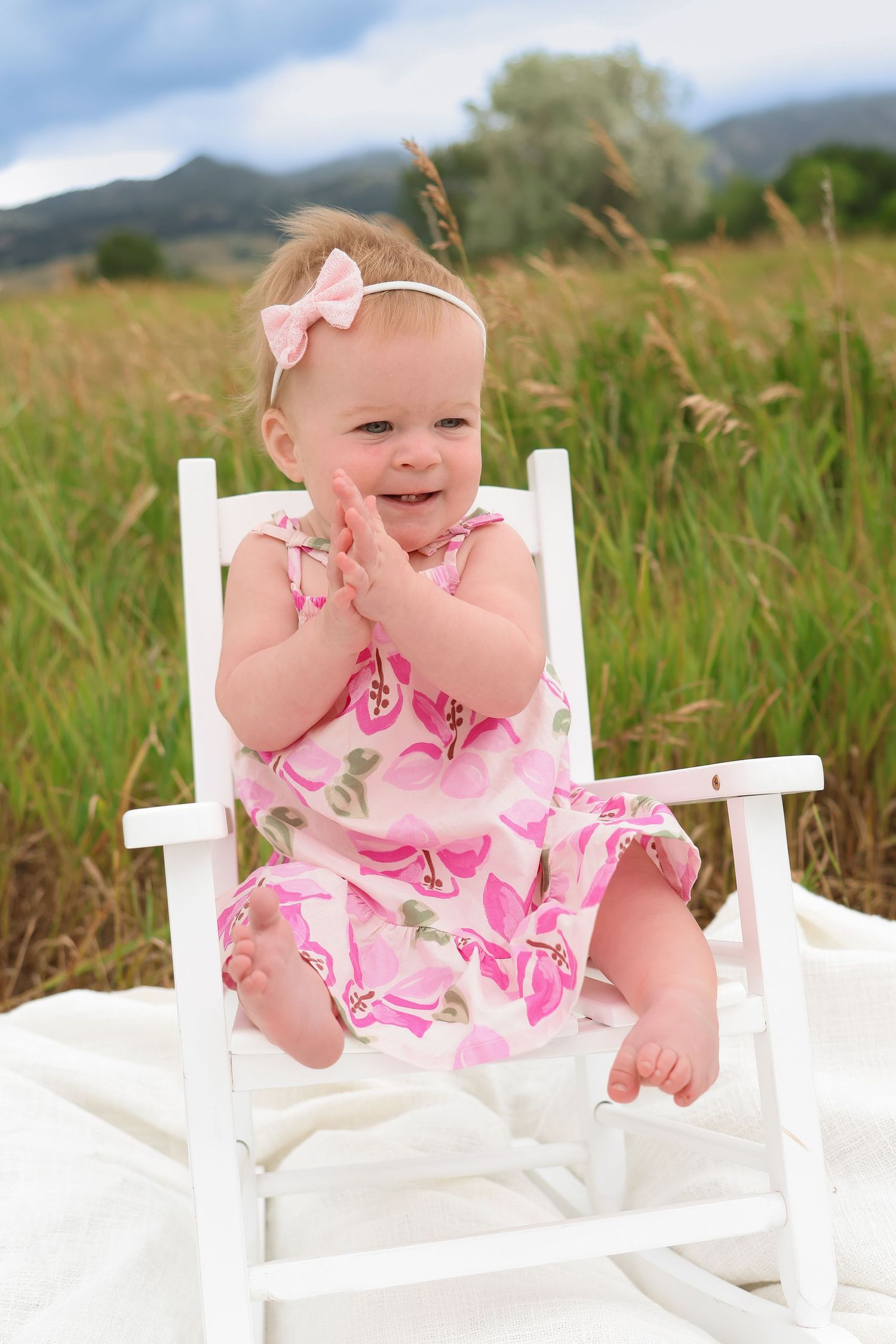Baby wearing pink floral dress and bow sits in white rocking chair outdoors in grassy field with mountains behind.