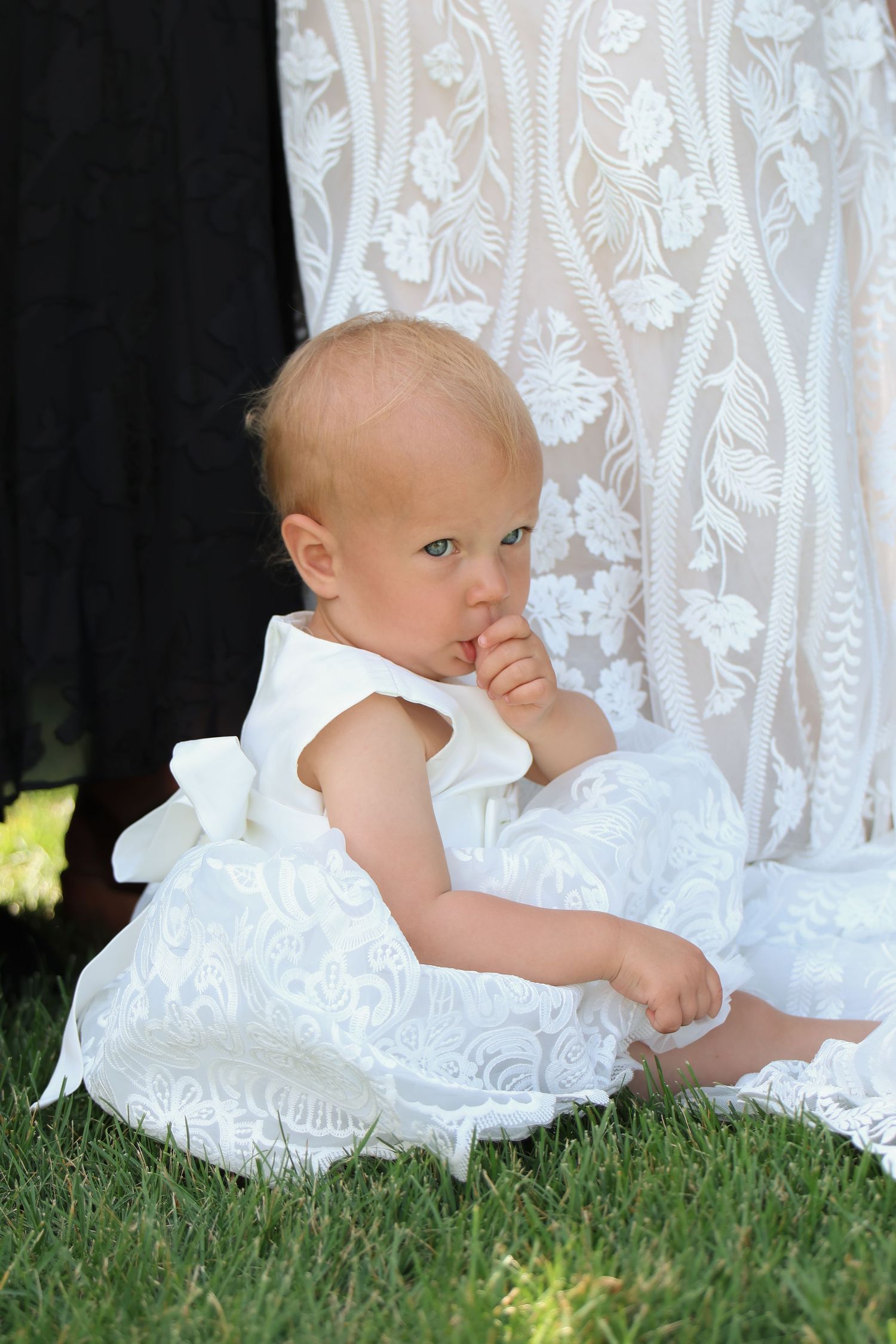 A baby in a white lace dress sits on grass beside her mom in a wedding dress while sucking their thumb.