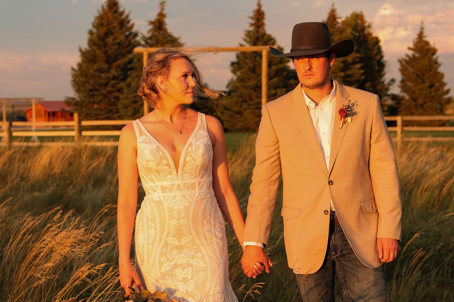 A couple holds hands in golden wheat fields at sunset, with bride in lace dress and groom wearing tan suit and cowboy hat.