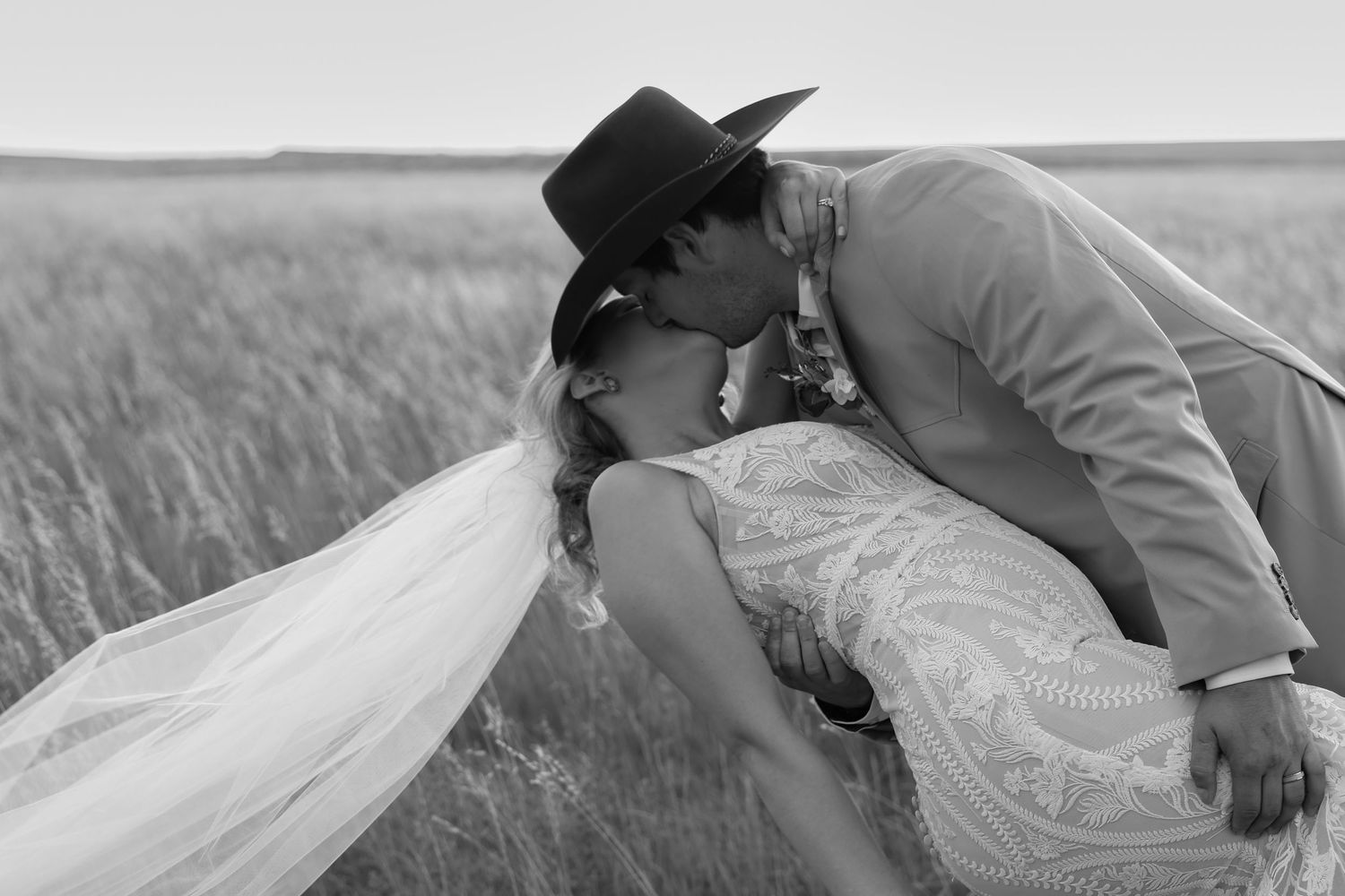 A romantic black and white wedding photo showing a couple kissing in a grassy field, with her veil flowing in the wind.