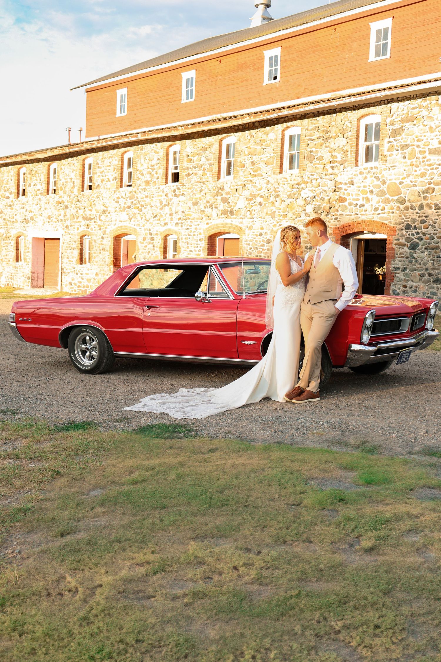 A bride and groom lean against a classic red car outside a rustic barn at sunset.