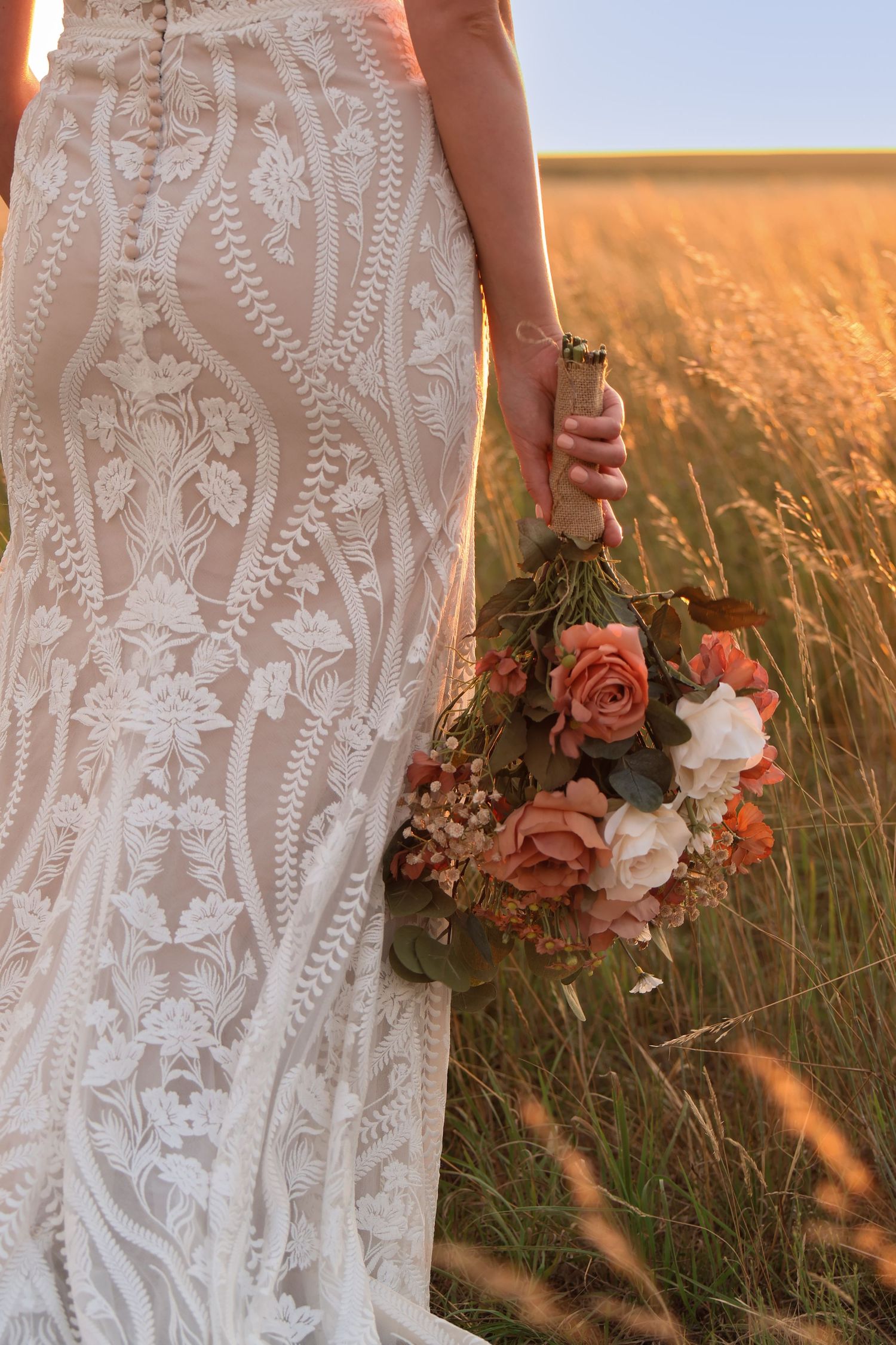 Elegant lace wedding dress details shown against golden wheat field holding coral and blush floral bouquet at sunset.