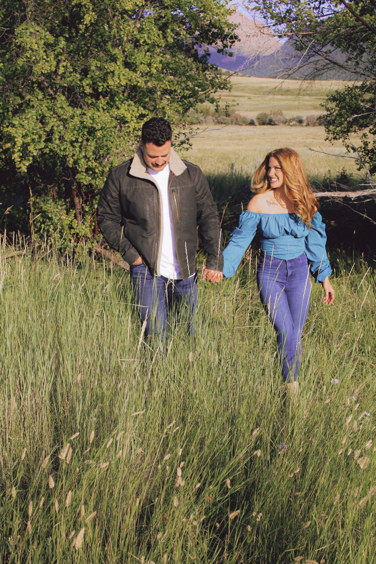Two people exploring together through a field of tall green grass on a sunny summer day between trees.