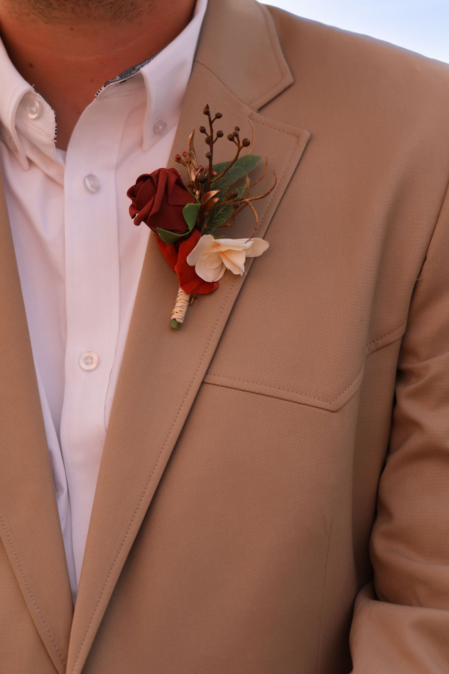 A burgundy and white floral boutonniere pinned to the lapel of a beige suit jacket with a white dress shirt.