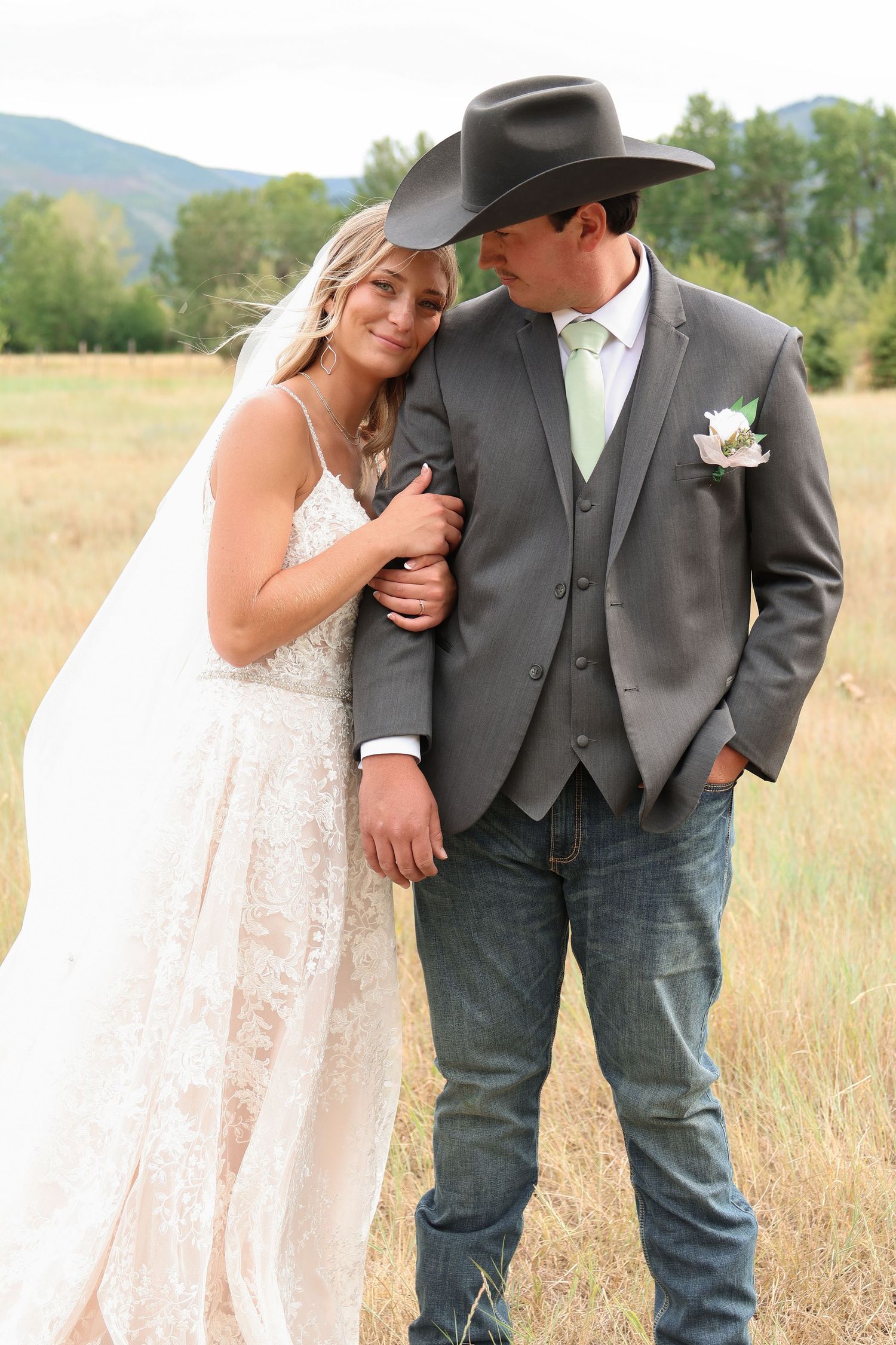 A country-western themed newlywed couple embraces in a scenic field with mountains in the background.