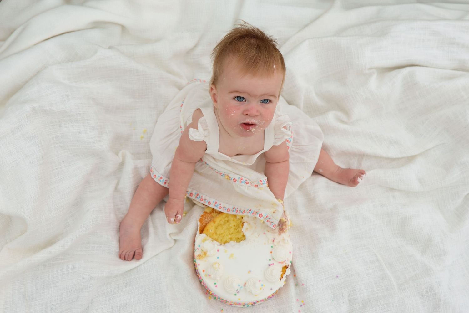 Baby wearing white ruffled dress sits on white fabric with cake smeared on their dress.