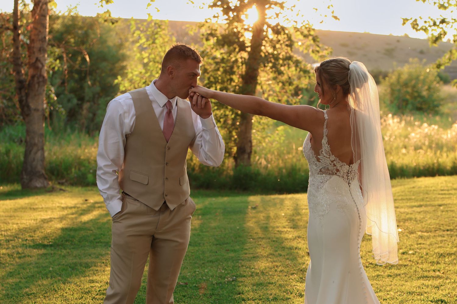 A couple shares a romantic golden hour moment as groom kisses her hand in a sunlit grassy field during their countryside wedding.