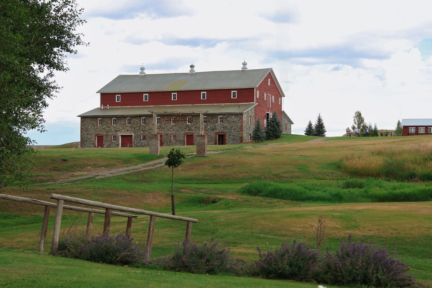 A rustic red barn with stone foundation sits atop a rolling green hill surrounded by countryside landscape in Helena, Montana.