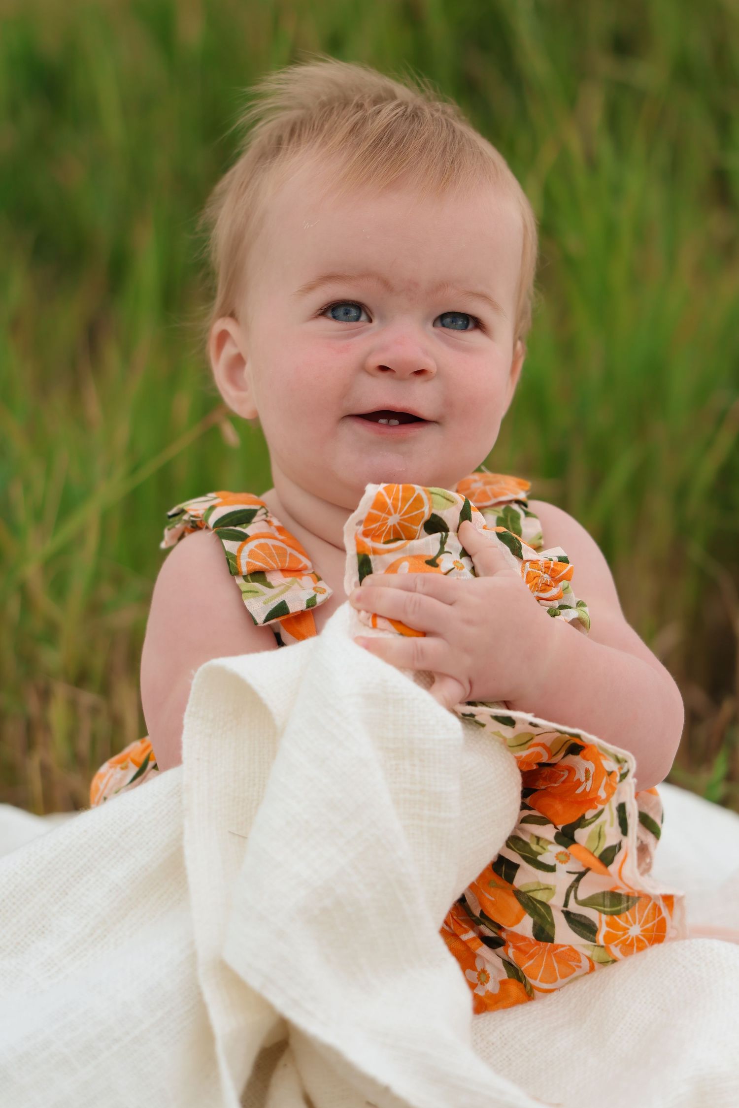 Sweet baby wearing orange floral dress sits on white blanket against green grassy background.