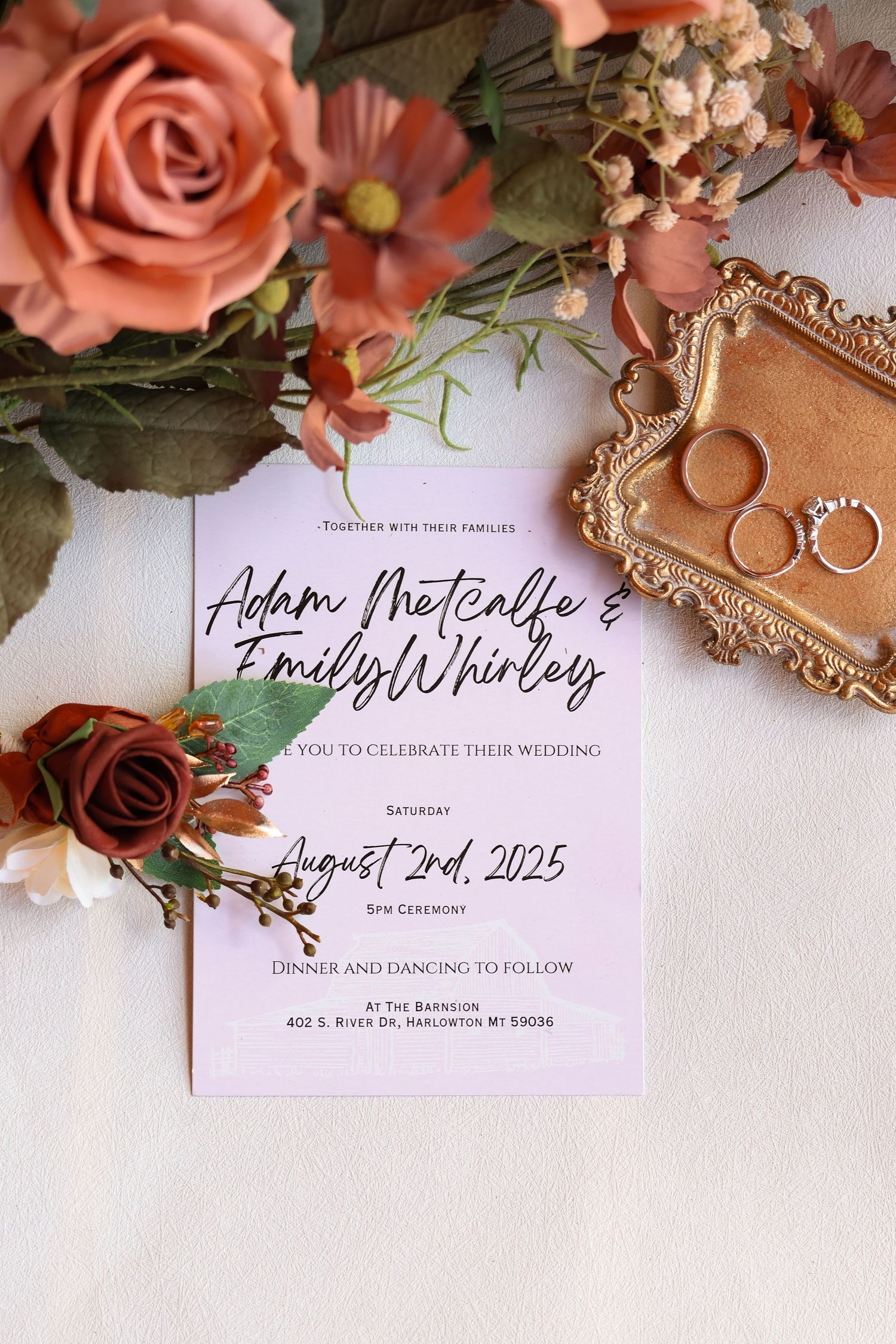 Wedding invitation with pink and red roses surrounded by wedding rings on an ornate gold tray against white backdrop.