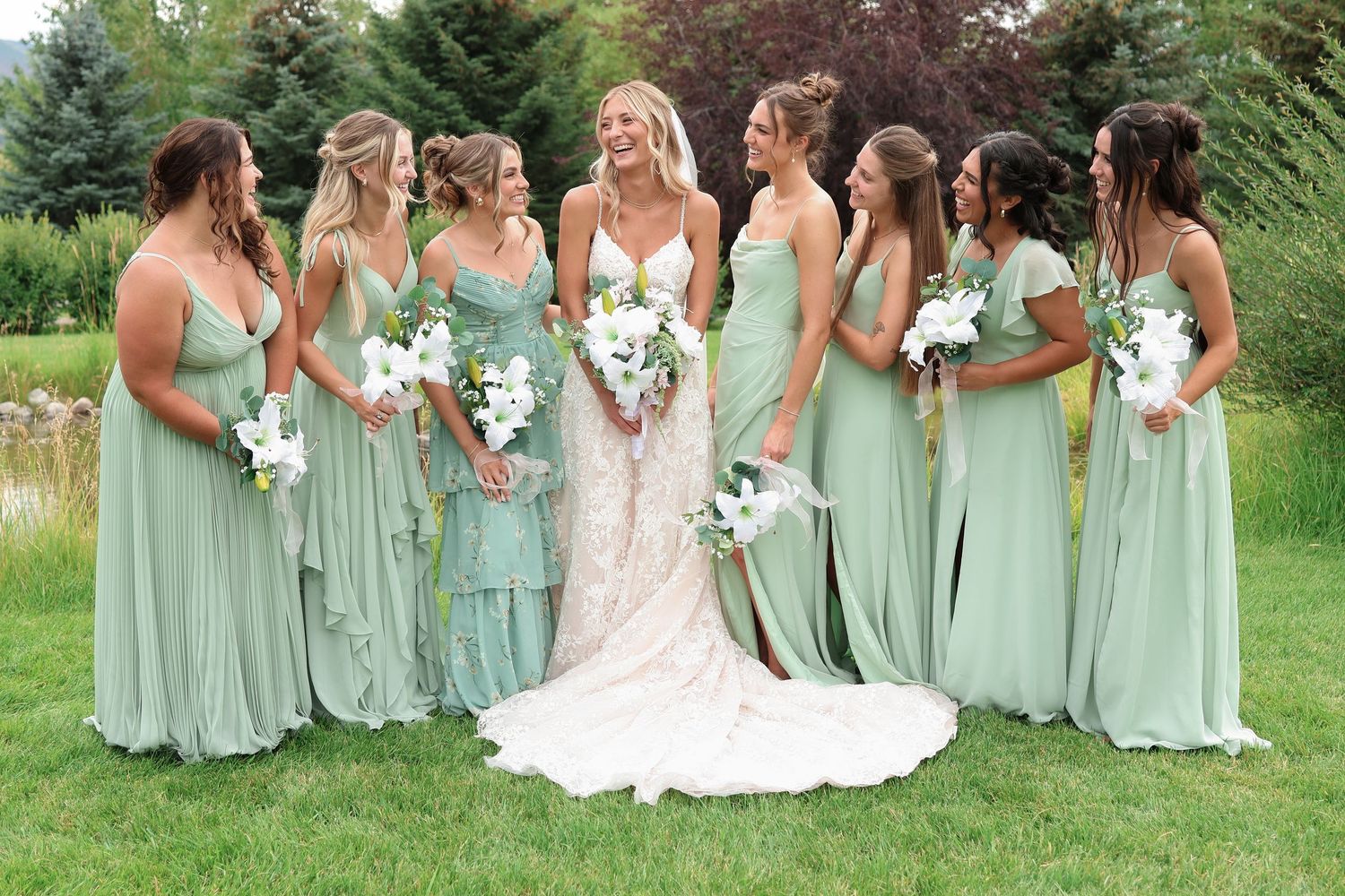 Wedding party wearing sage green bridesmaid dresses gathering on lawn holding white floral bouquets.