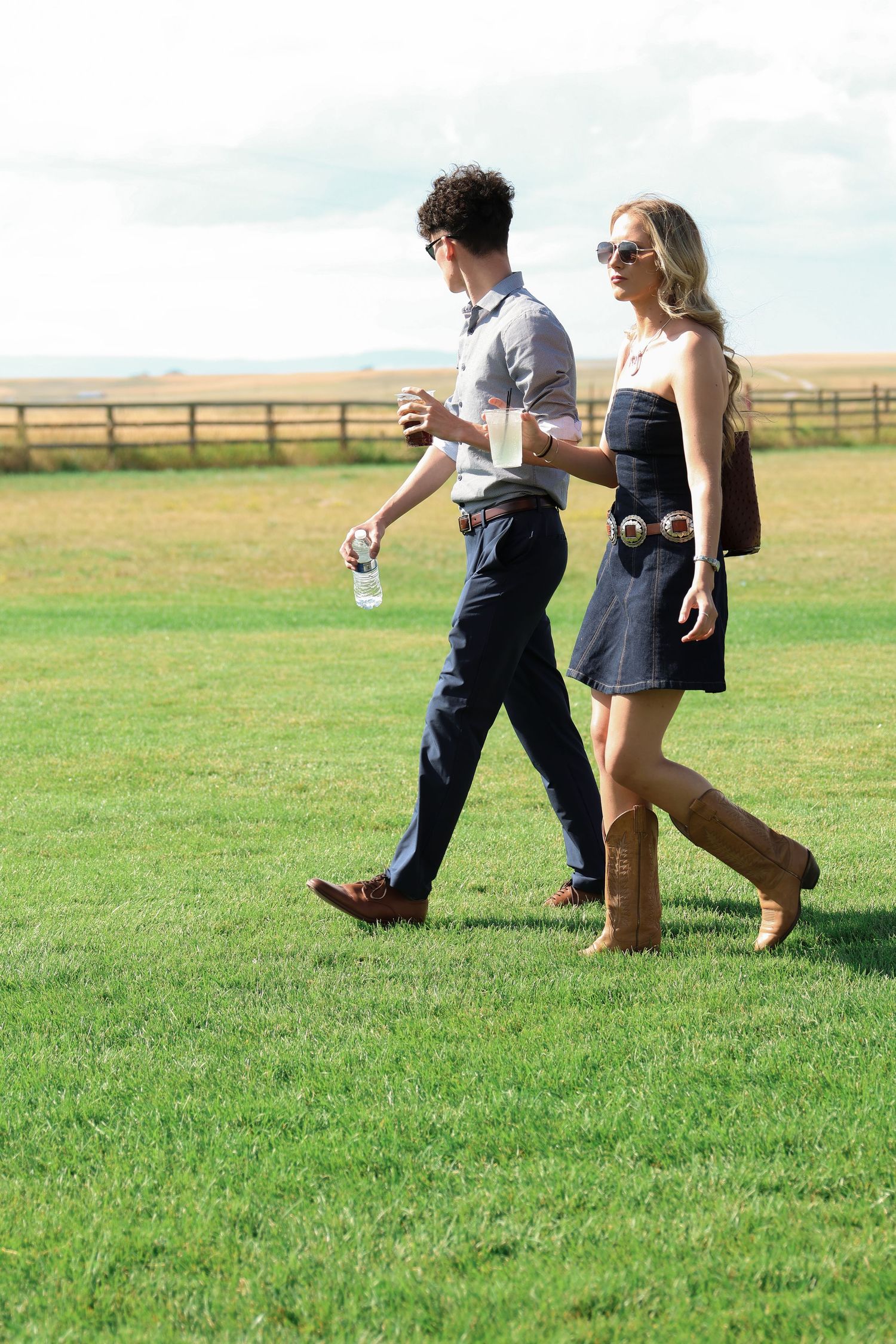 Western chic couple walking together on lush green grass with wooden fence and countryside views in the background.