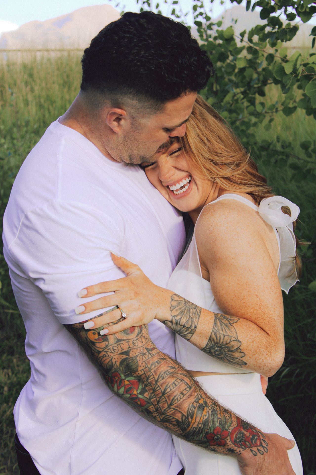 A tender embrace between a couple outdoors, with colorful tattoo sleeves and white attire against a natural backdrop.
