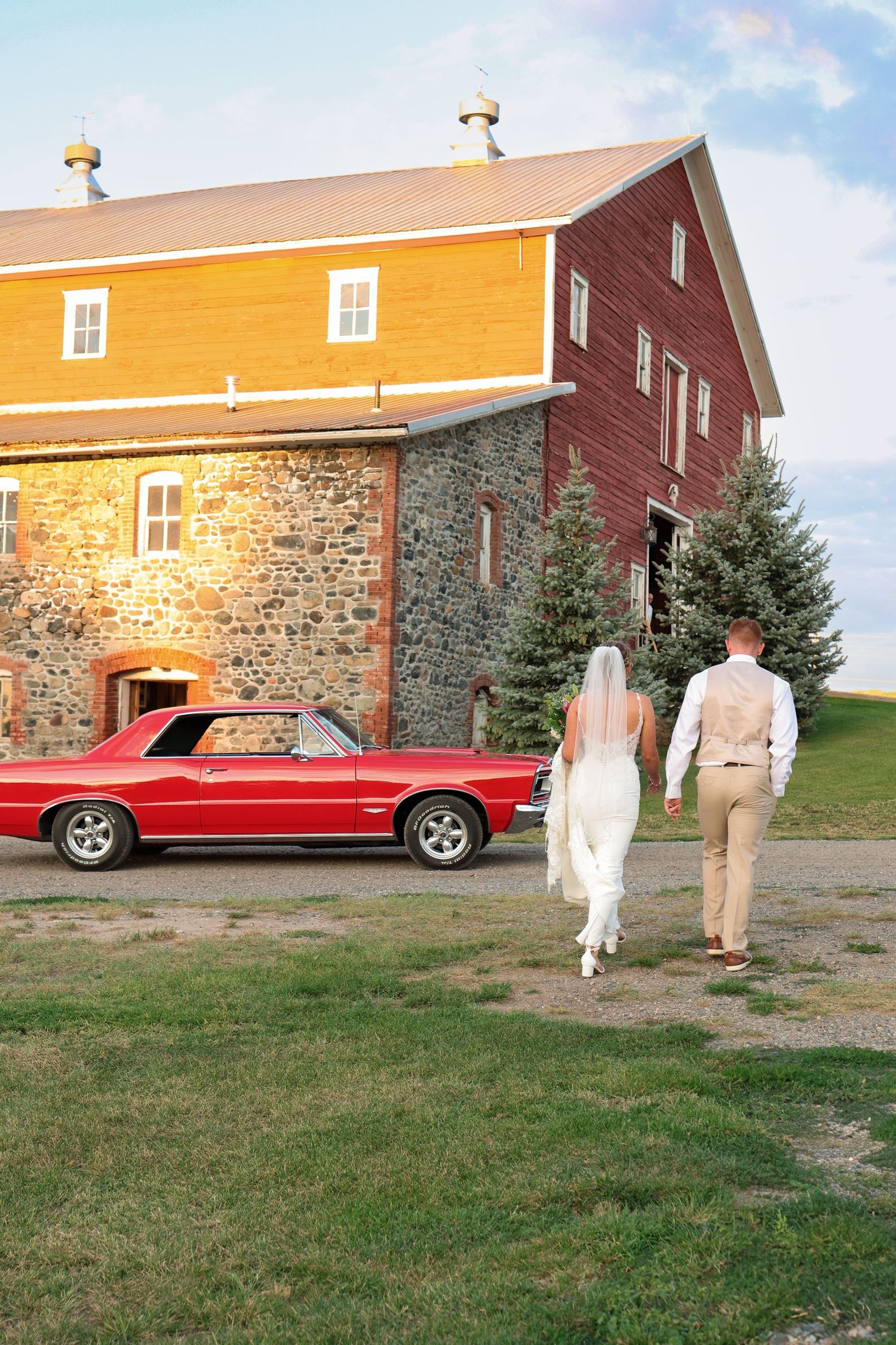 A bride and groom walks towards a rustic stone and wood barn with a vintage red Mustang parked outside during golden hour.