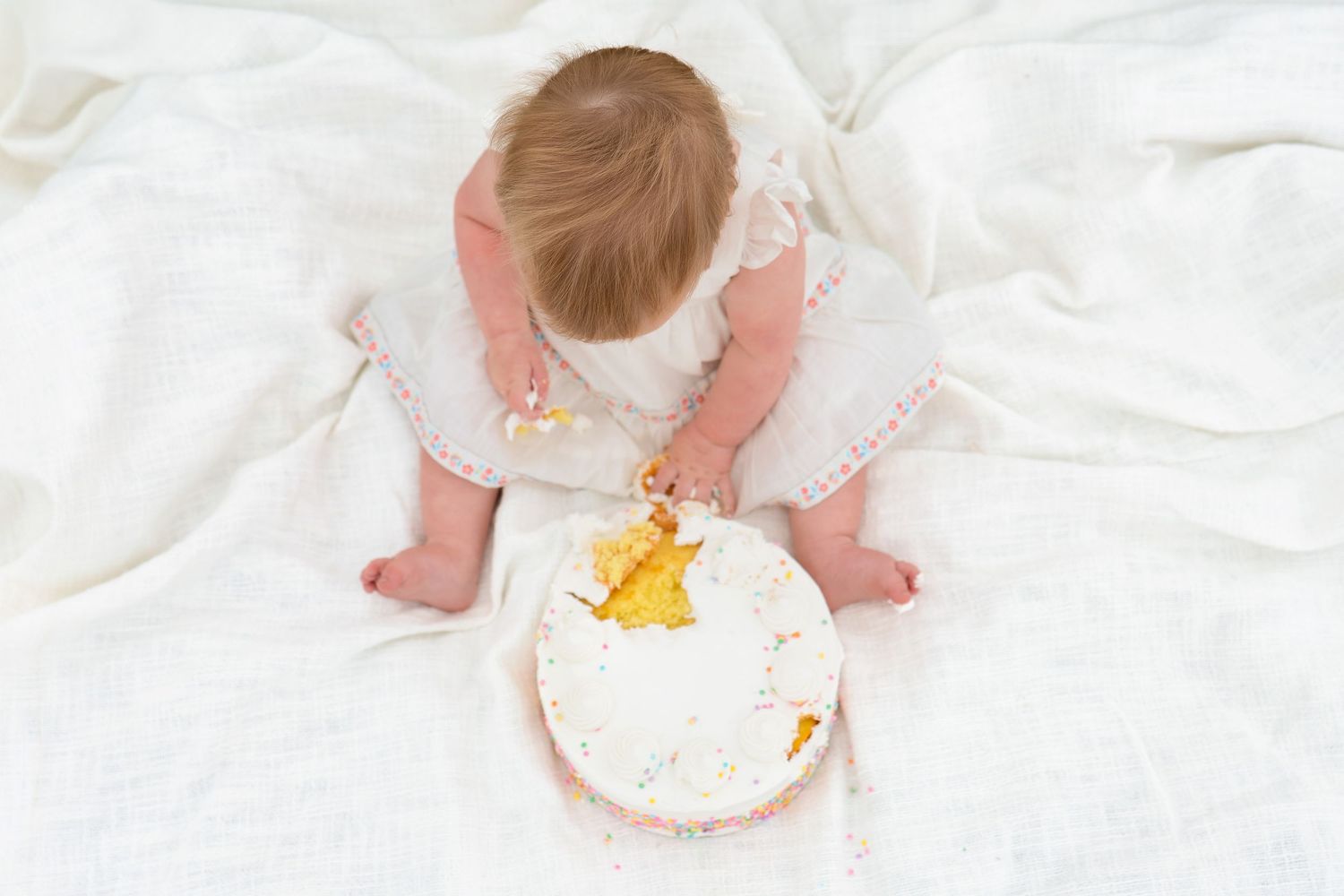 A small baby in white clothes sits on a white blanket looking down at a birthday cake with a yellow number one candle.
