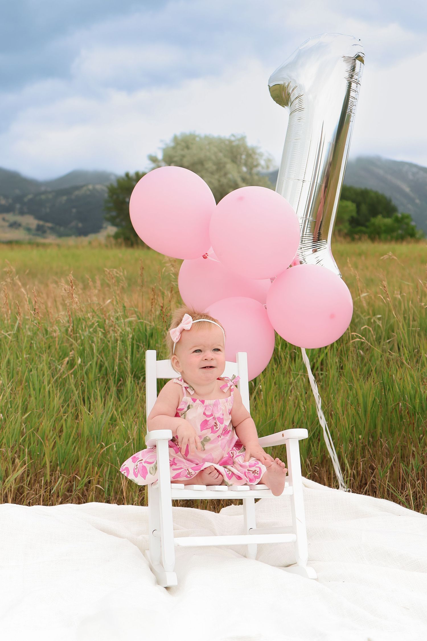 First Birthday celebration setup with pink balloons and white chair against mountain landscape backdrop.