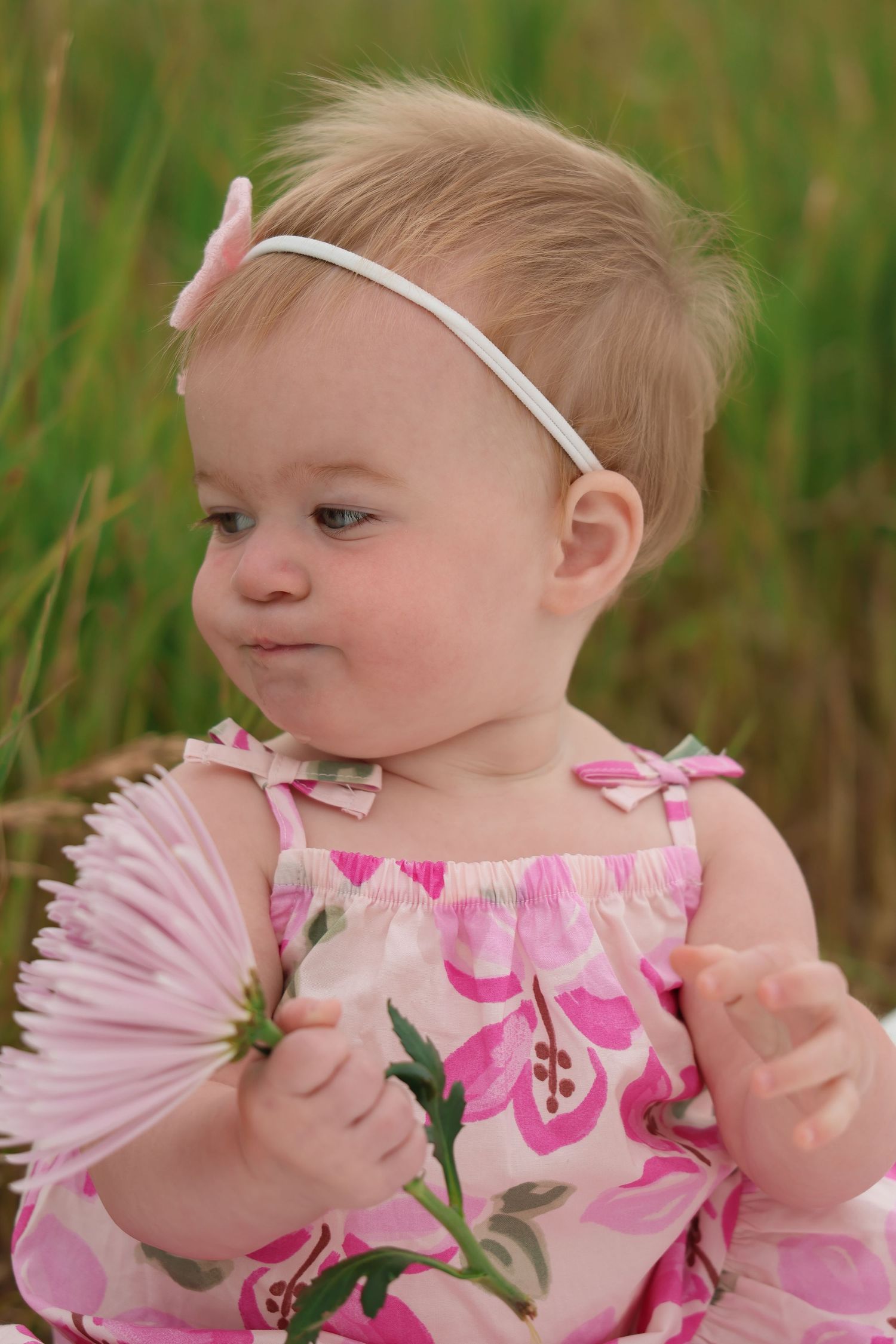 Baby wearing pink floral sundress and white headband holds a pink flower while sitting in grassy field.