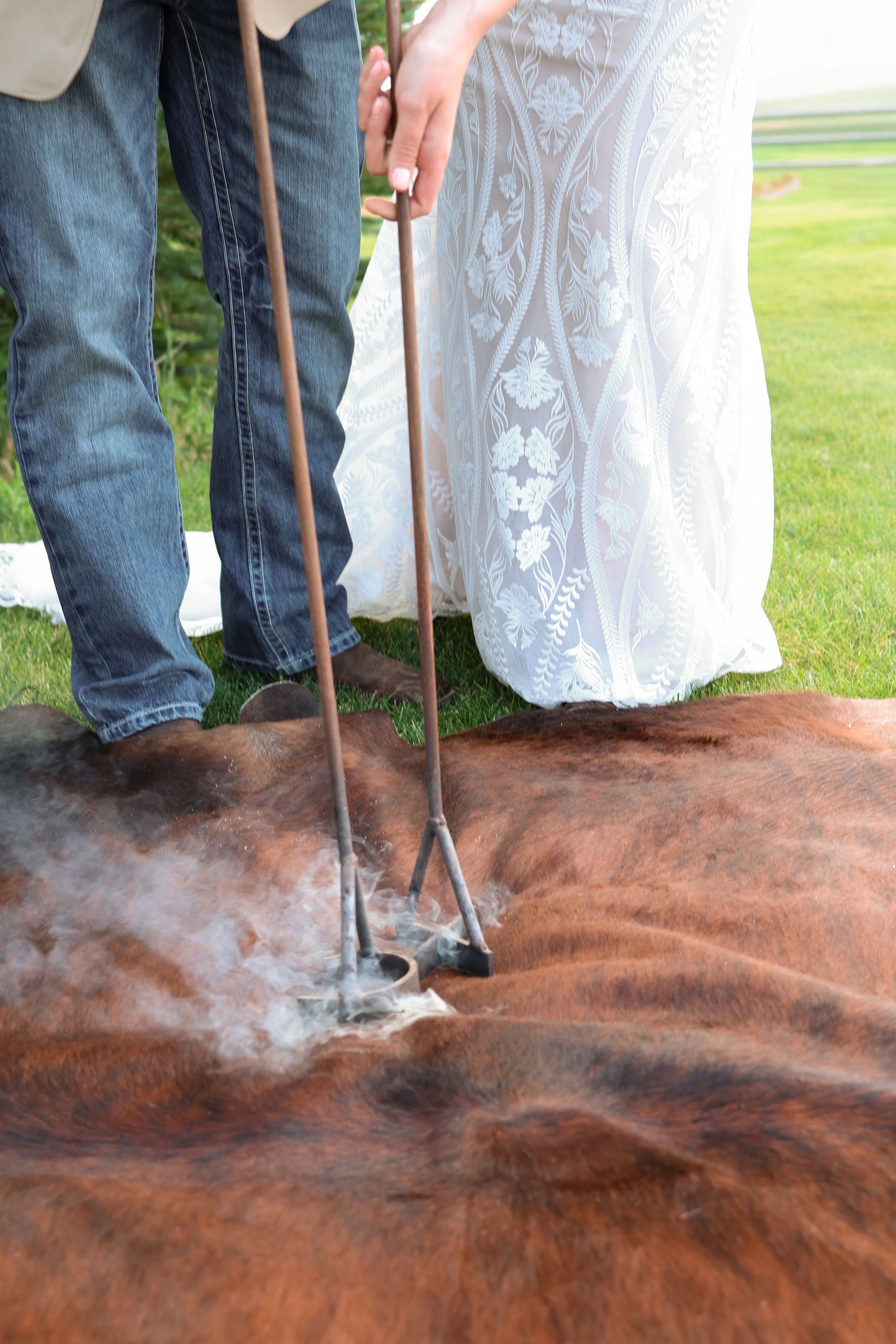 Intimate branding of cow hide with hot irons while wearing blue jeans and a white lace wedding dress on grassy field.