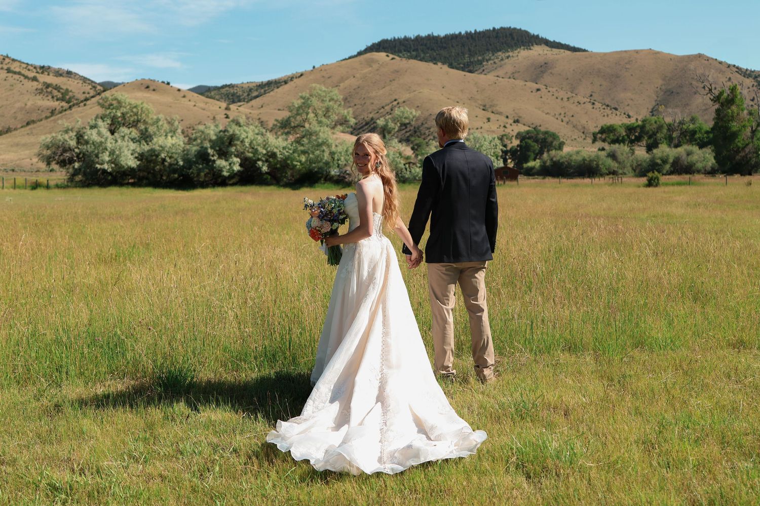 A couple in wedding attire walks through a golden meadow with Montana's rolling hills and mountains in the background.
