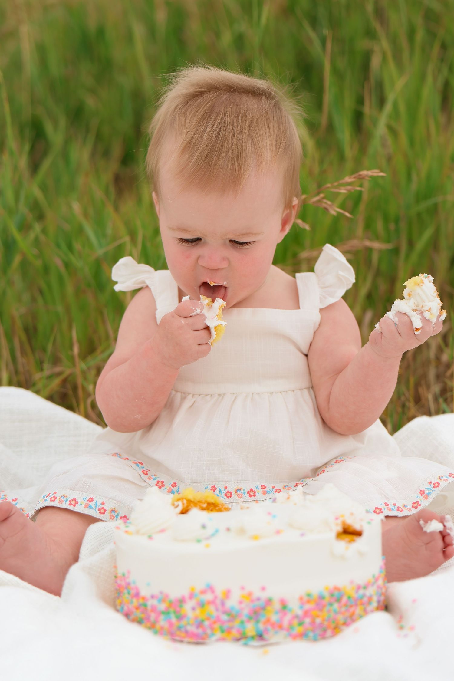 A baby in a white dress enjoys a birthday cake with sprinkles while sitting on a white blanket in tall grass.