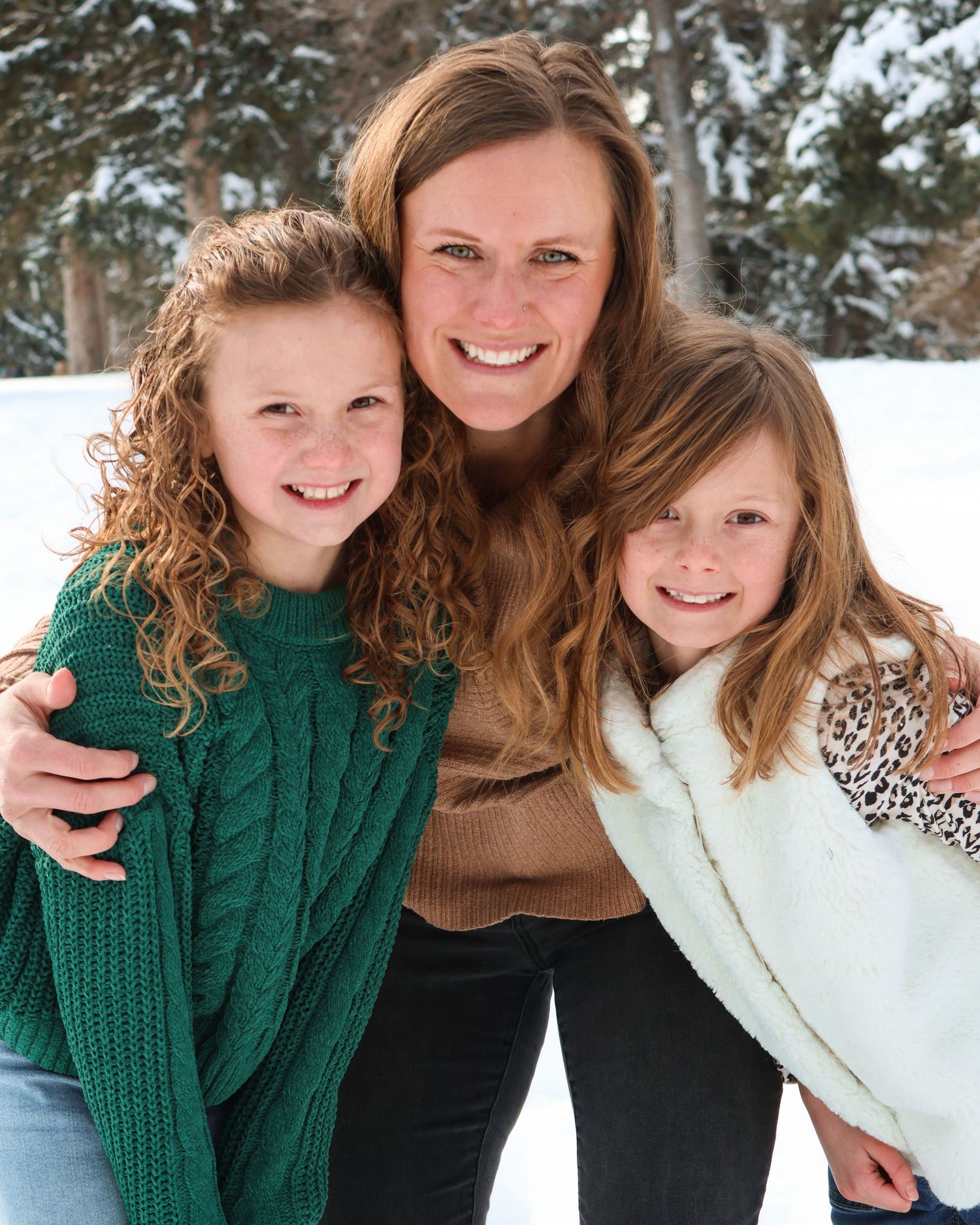 A heartwarming winter portrait of a Mom and her daughters in cozy sweaters smiling together against a snowy background.