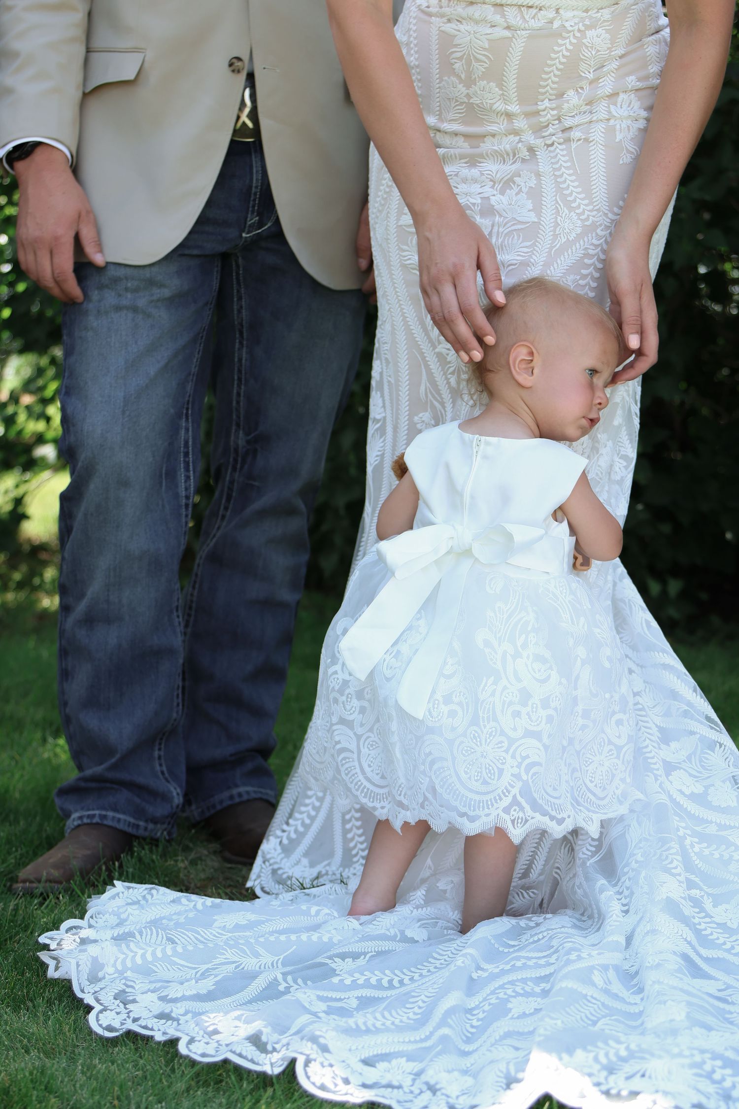 A small child in a white dress stands between parents in wedding attire on grass, with lace details.