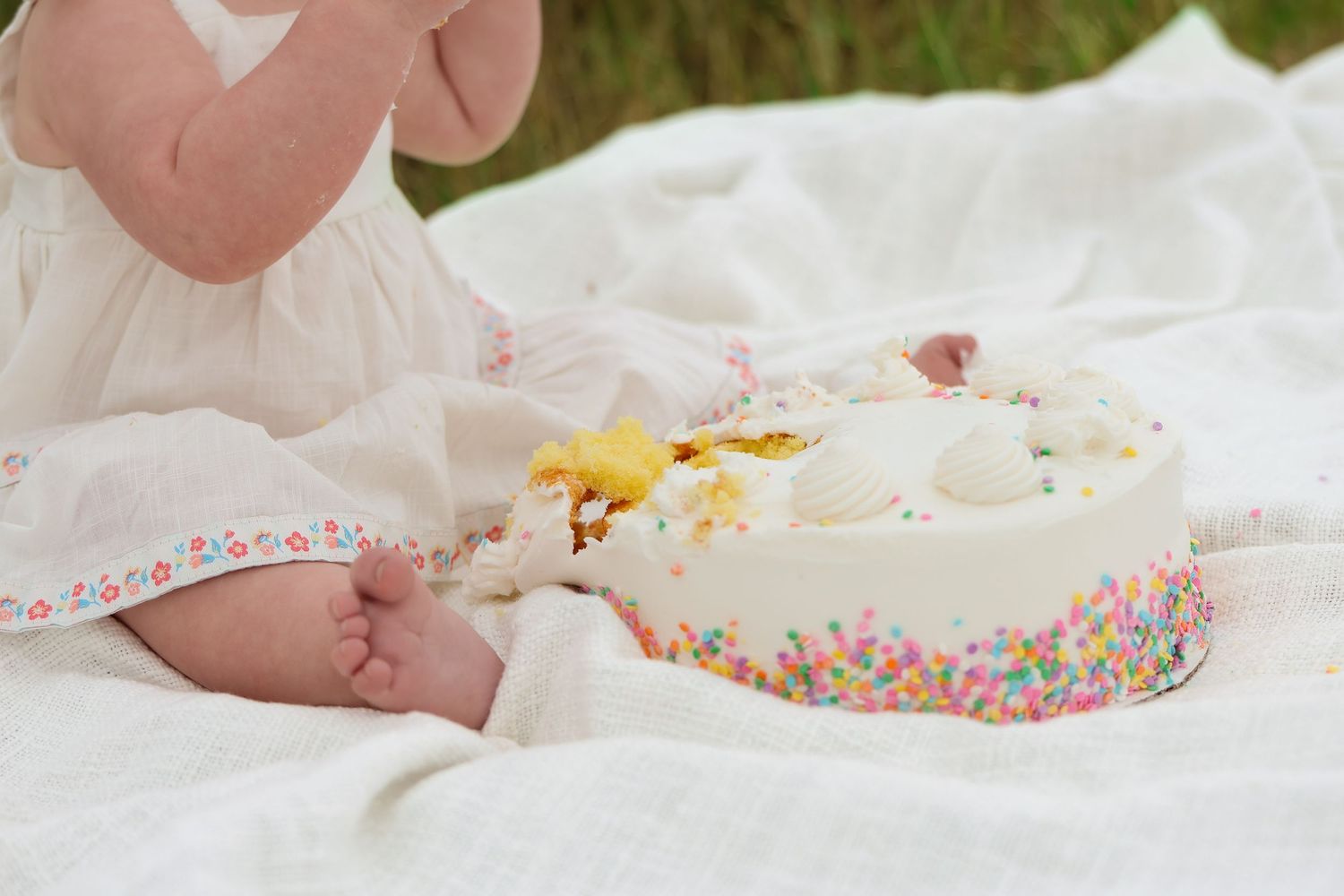 Small feet rest on white blanket beside birthday cake decorated with sprinkles and frosting at outdoor picnic setup.