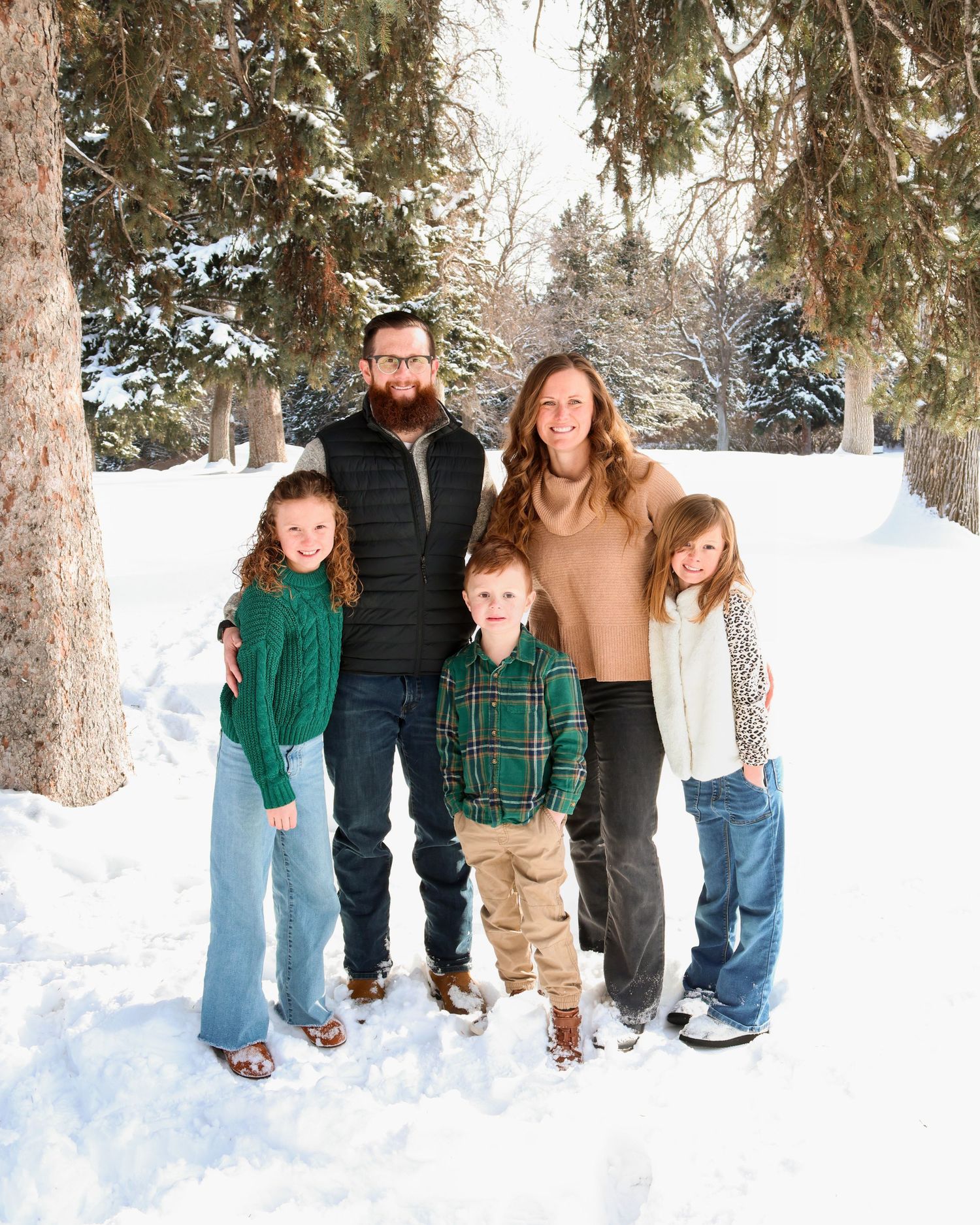A family stands together outdoors in the snow surrounded by evergreen trees during a winter photoshoot.