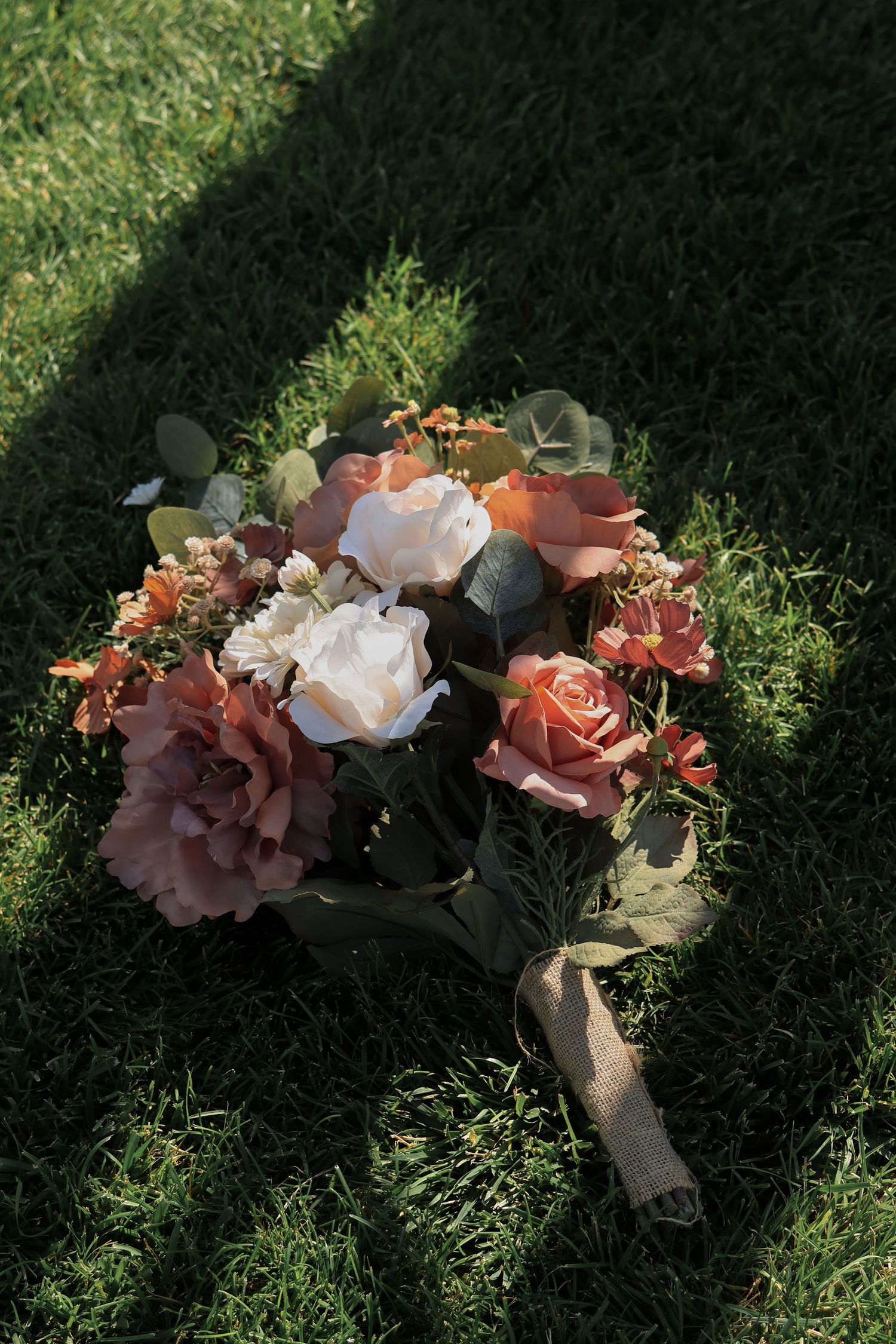 A romantic bridal bouquet with orange, pink and white roses laying on grass in natural sunlight with cast shadows.