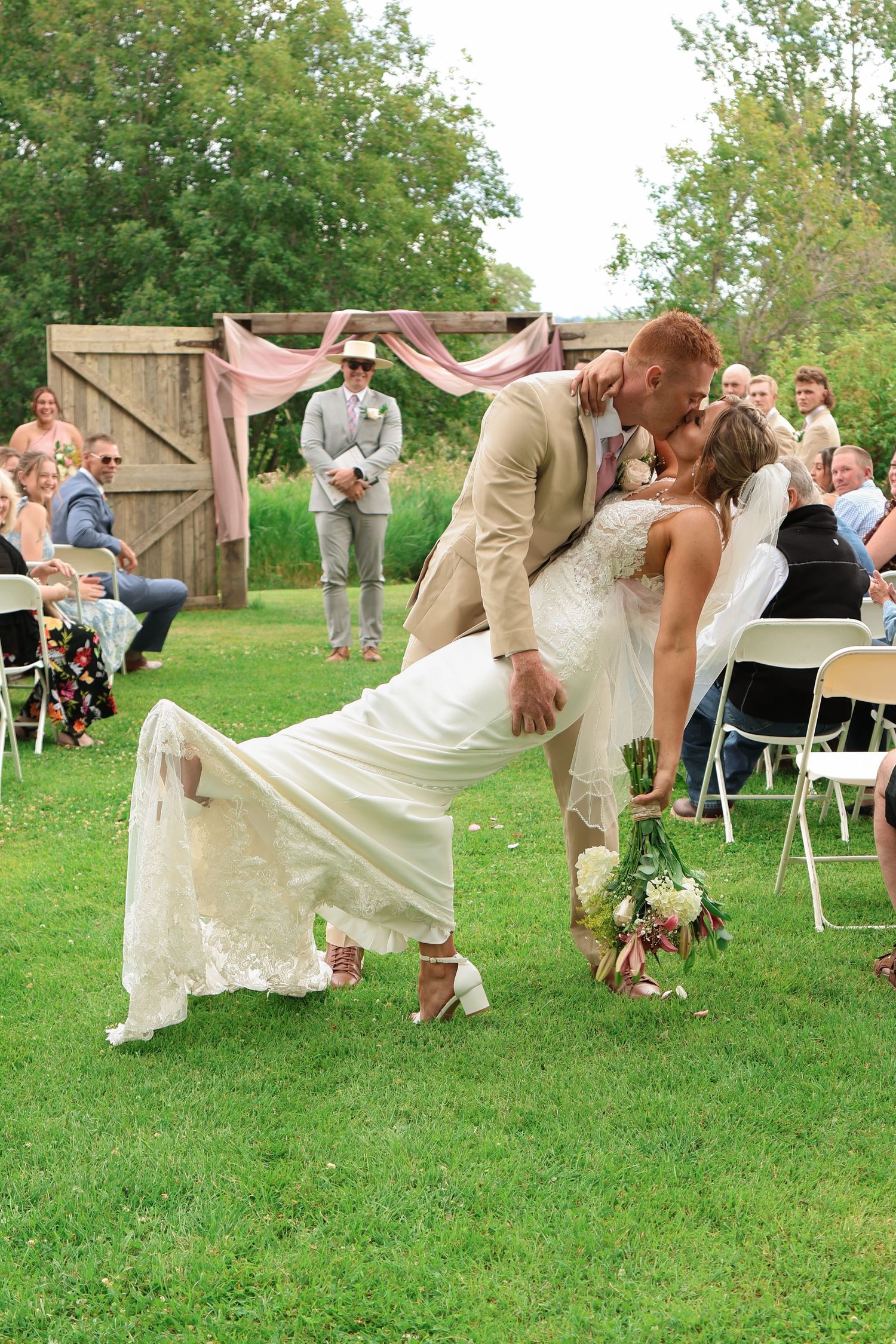 Couple shares romantic dip kiss during outdoor garden wedding ceremony with wooden arch and pink drapery.
