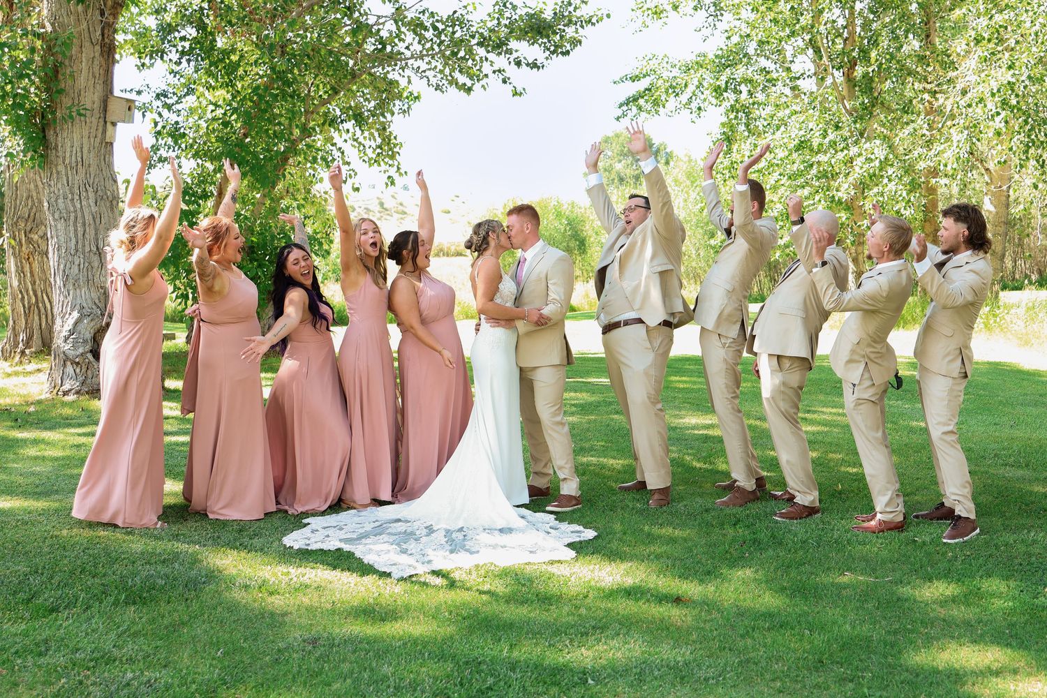 Wedding party wearing dusty pink dresses and tan suits poses playfully in a row along trees on a grassy lawn.
