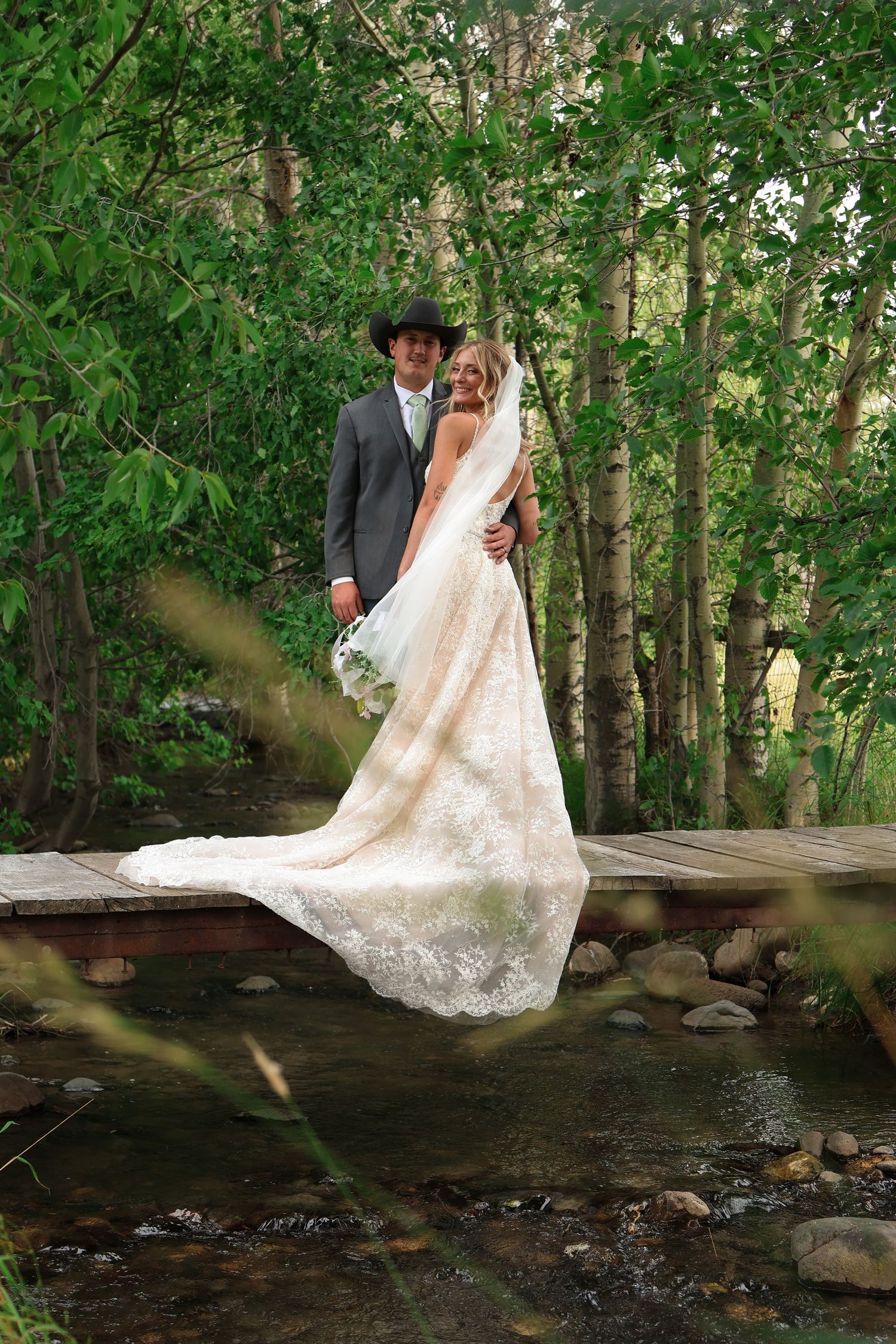 A couple in wedding attire pose together on a wooden bridge over a rocky creek in a lush forest setting.