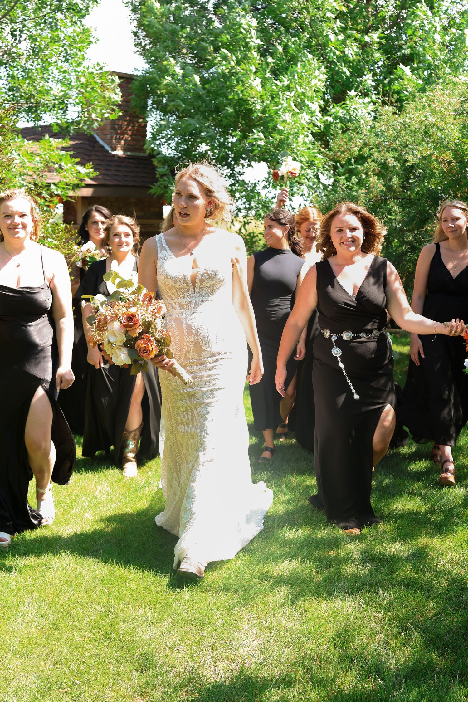 Wedding party members in black dresses and the bride in white pose outdoors on a sunny day with green trees in background.